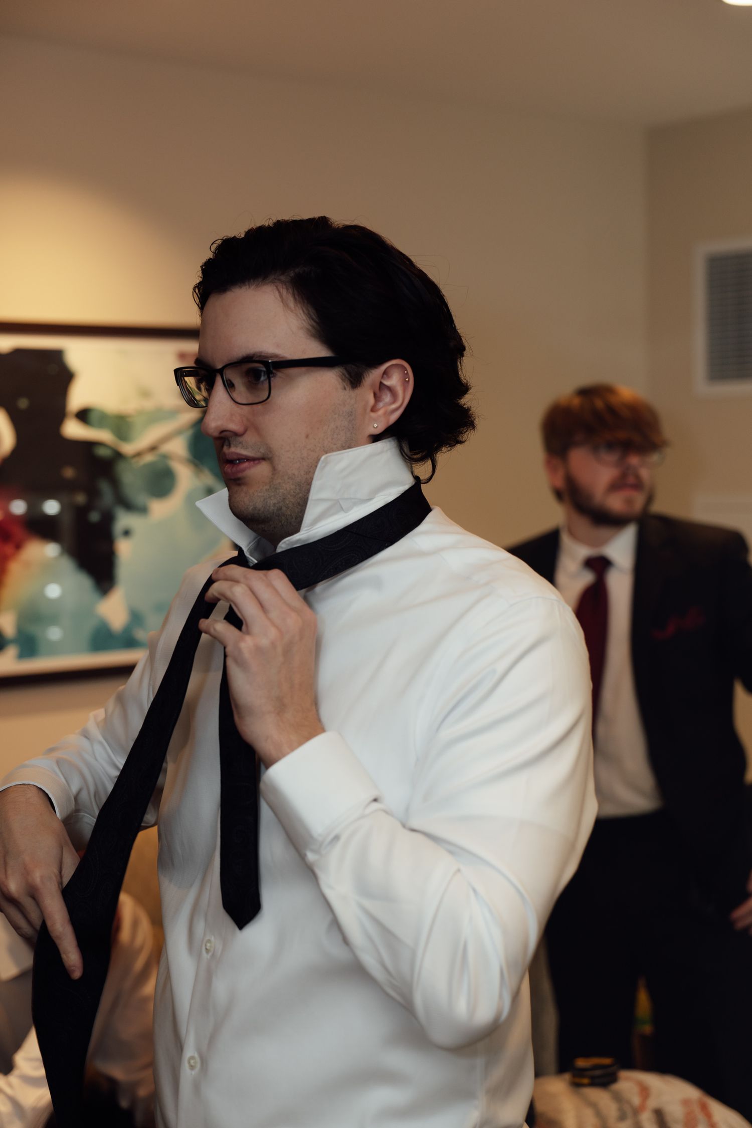 Man in white shirt adjusting black tie while getting dressed for formal event, with guest in background.