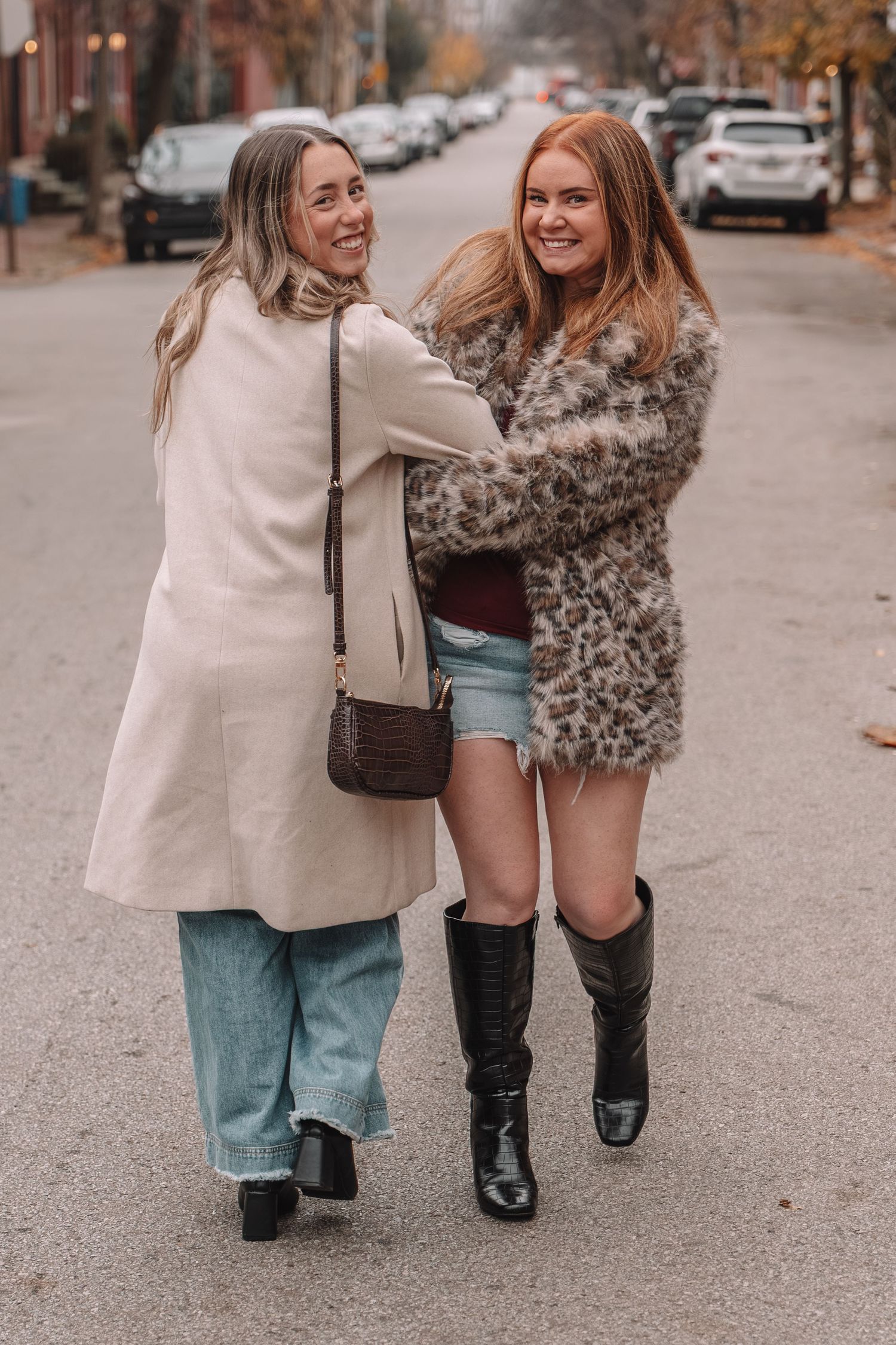 Two smiling women walking arm in arm on a city street, wearing stylish fall outfits including coats and boots.