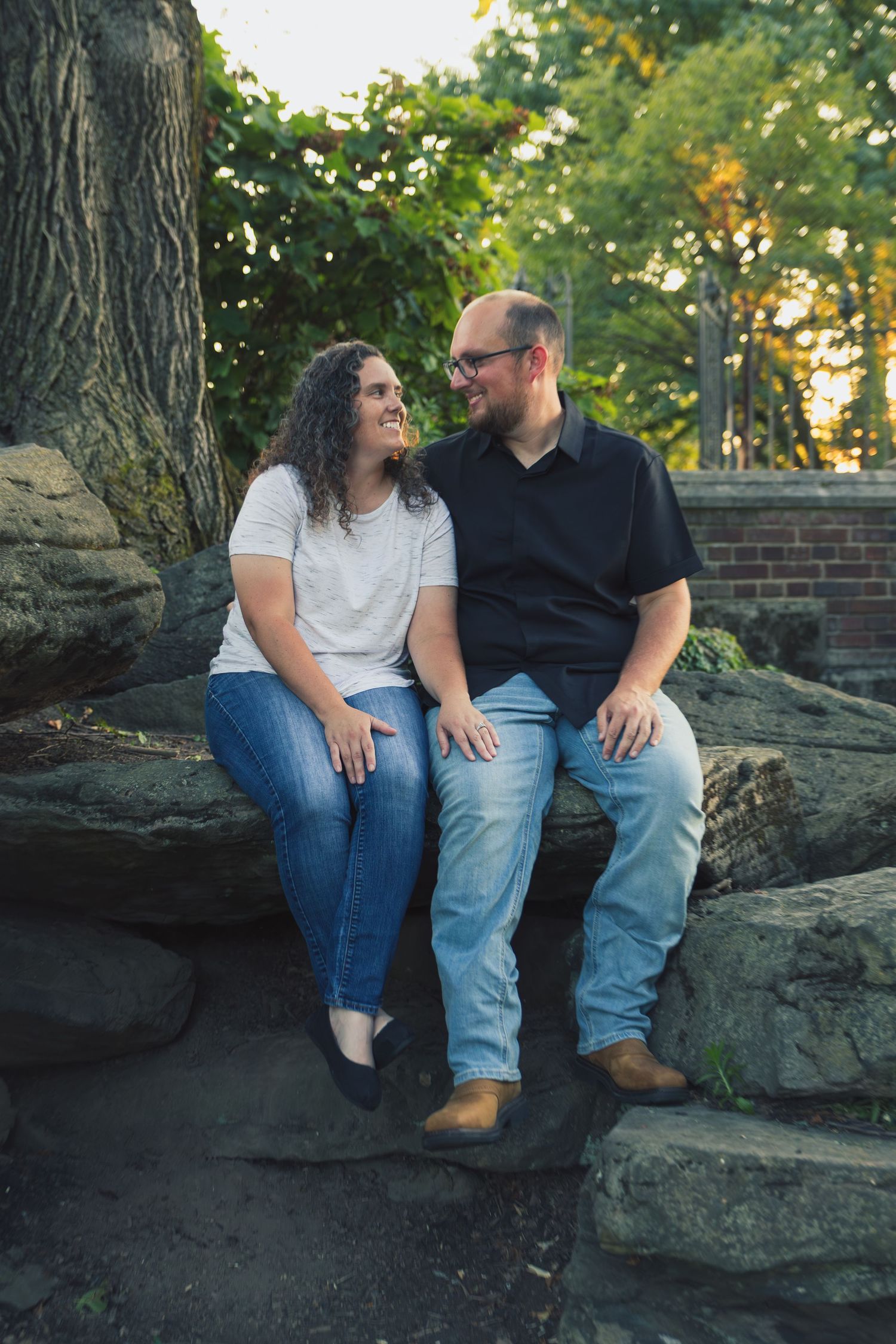 A smiling couple sits together on rocks in a park, the woman in a white top and the man in a black shirt, sharing a joyful moment.