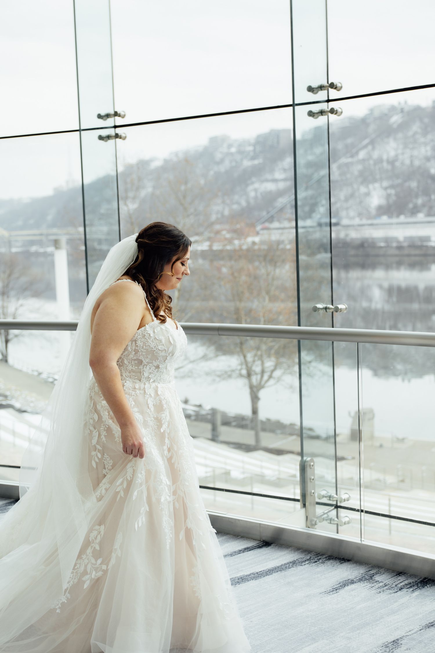 Bride in lace wedding gown and veil stands by floor-to-ceiling windows with snowy river view.