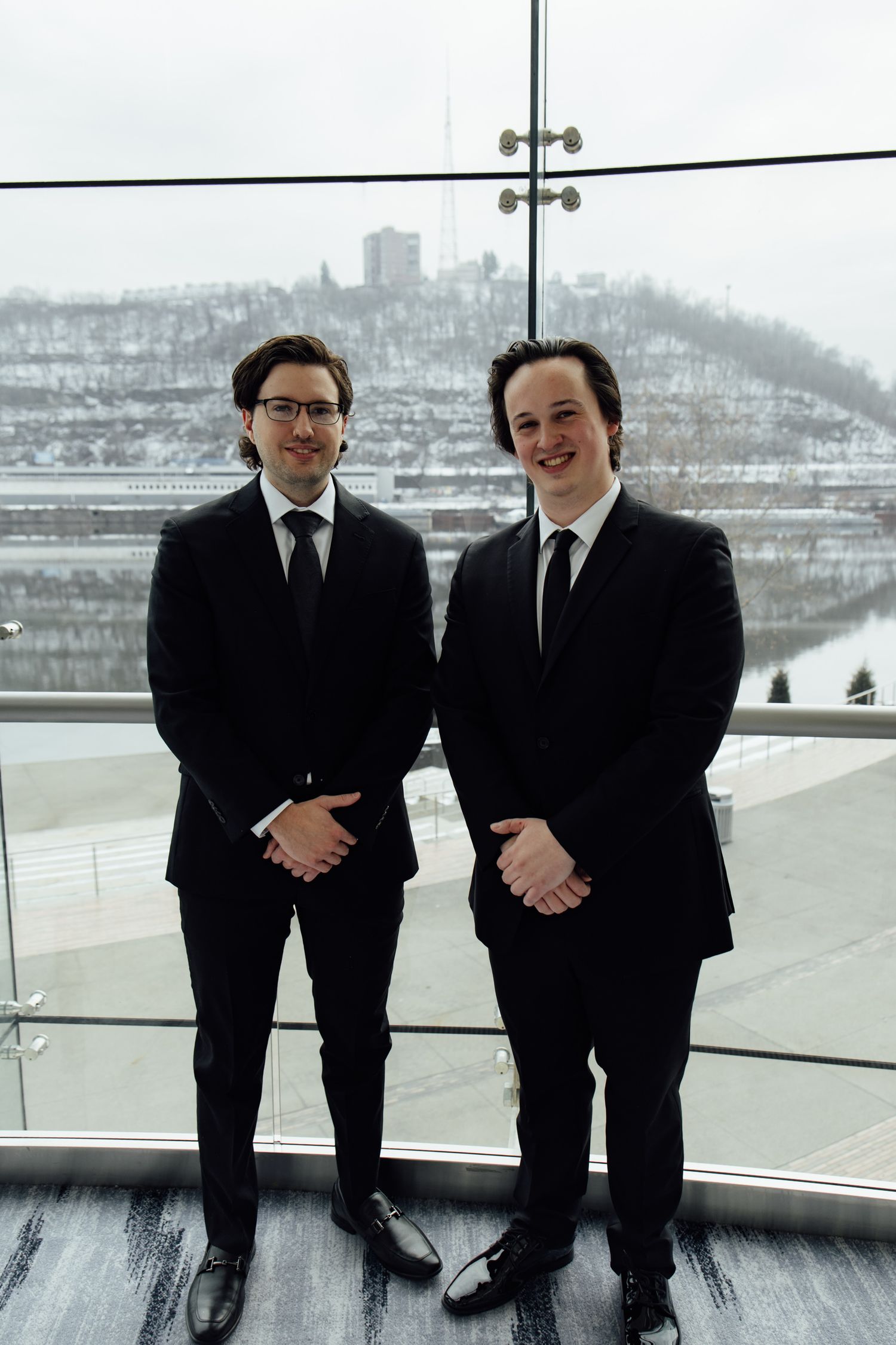 Two men in black suits and ties standing indoors with a snowy cityscape visible through large windows behind them.