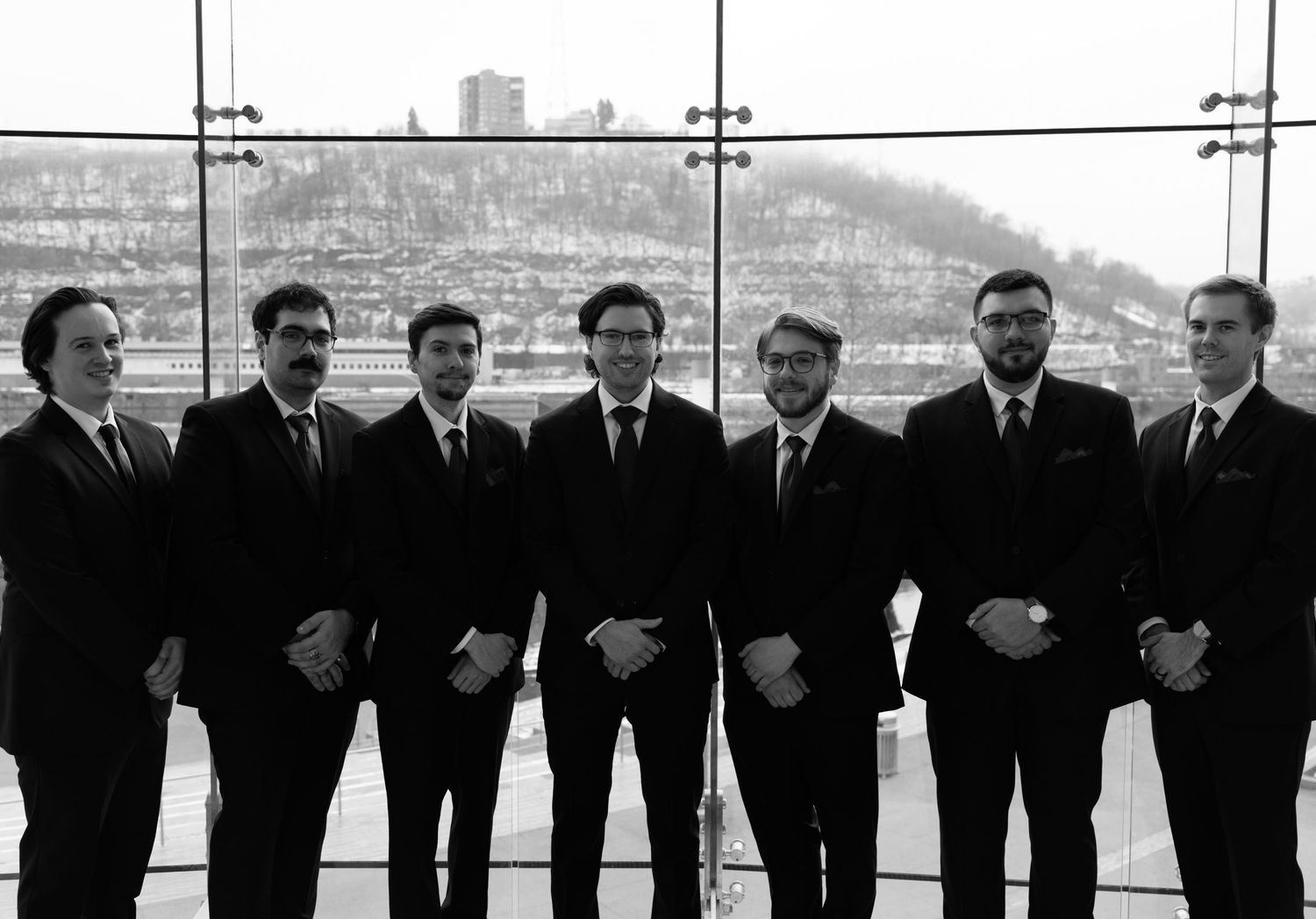 Seven men in dark suits pose together in a formal group photo with a scenic hillside view in the background.