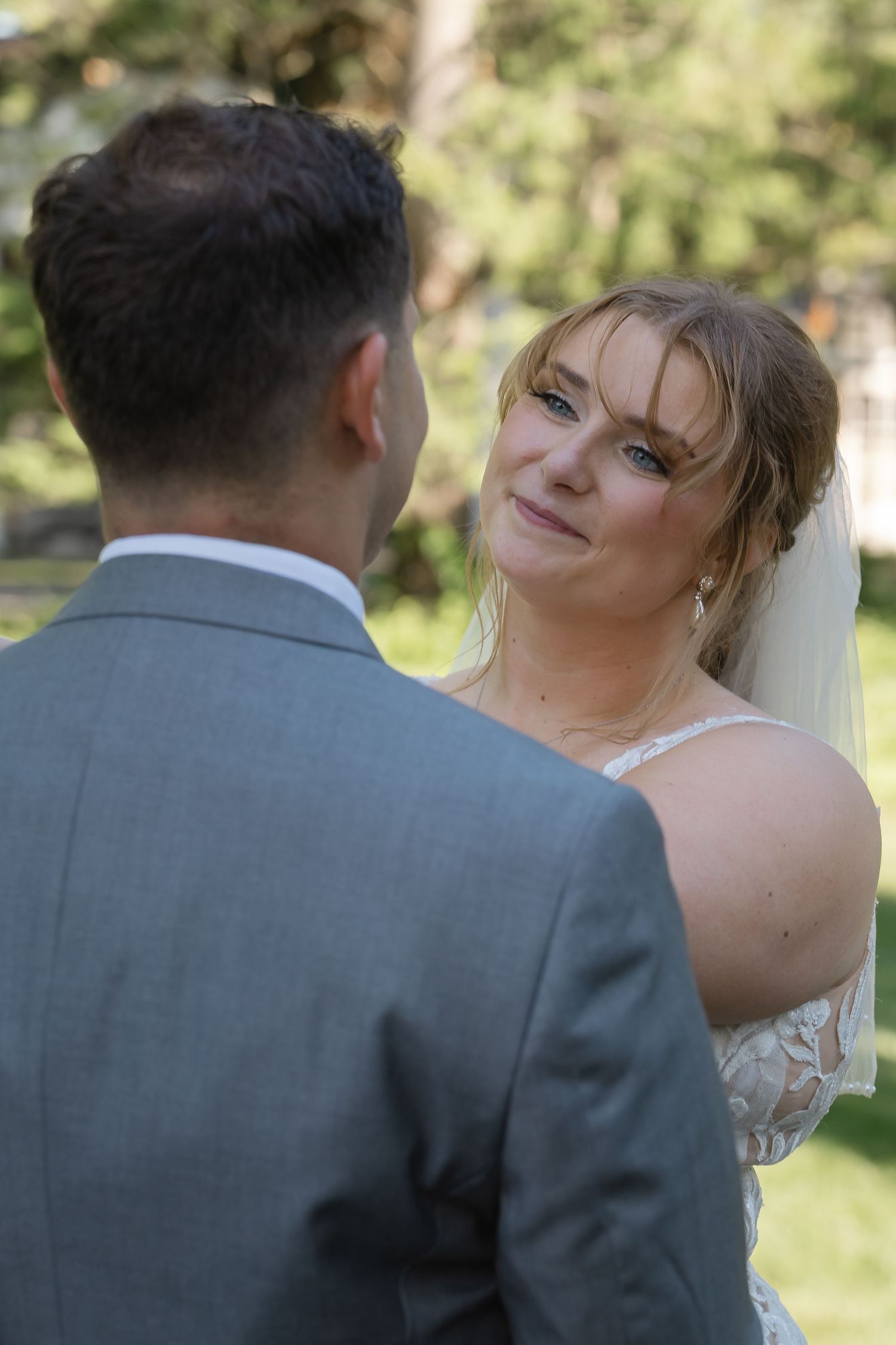 Bride in lace dress and veil smiles emotionally at groom in gray suit during outdoor wedding ceremony.