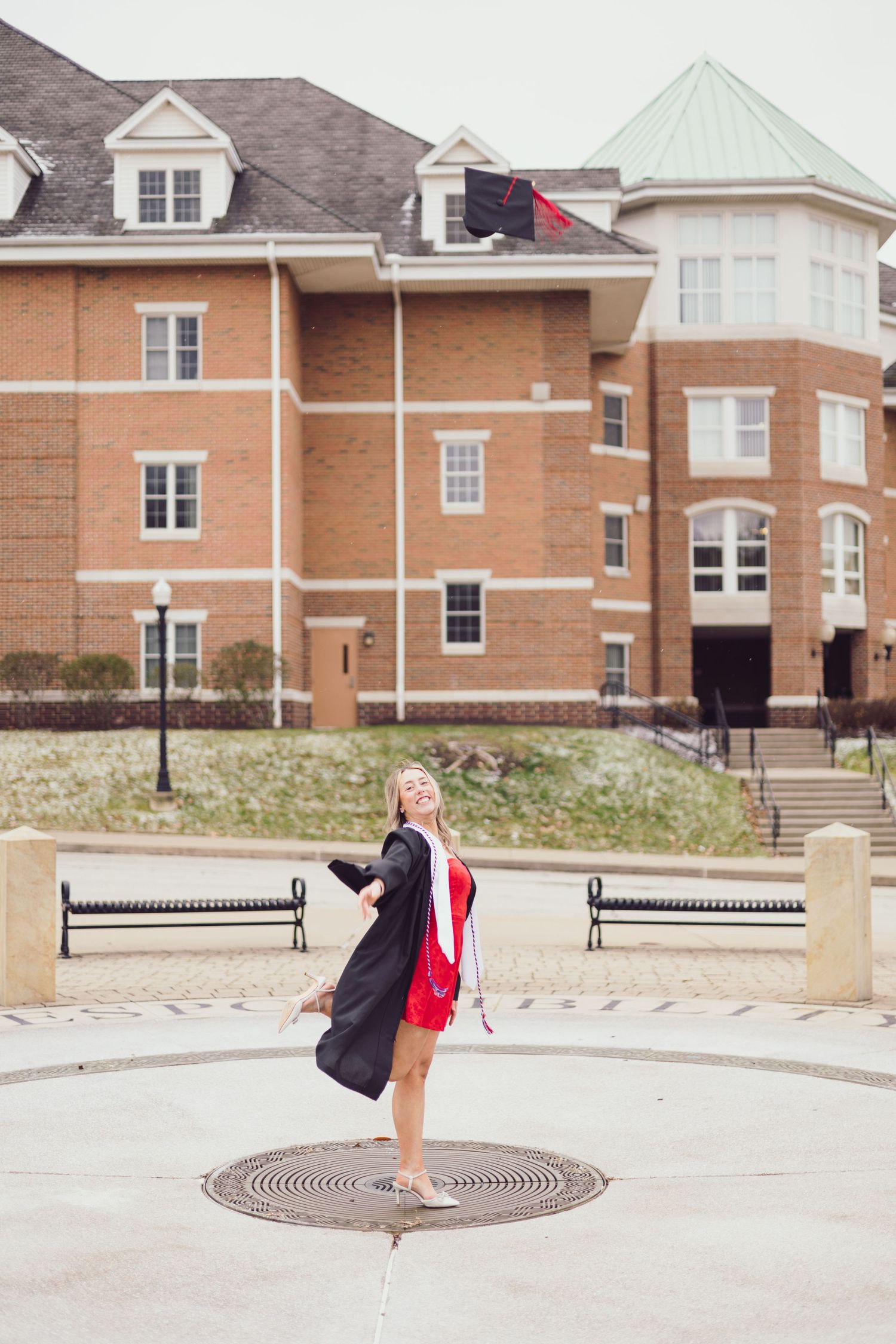 Graduate in cap and red dress twirling outside brick university building.