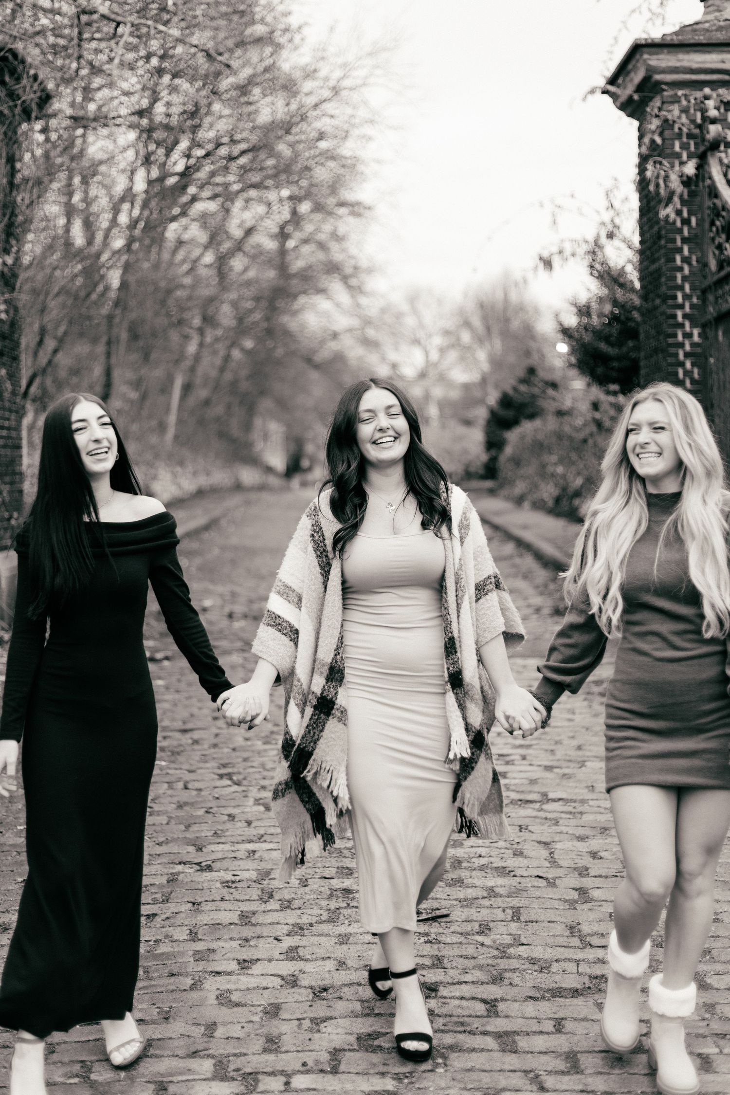 Black and white photo of three friends walking hand in hand on a cobblestone path.