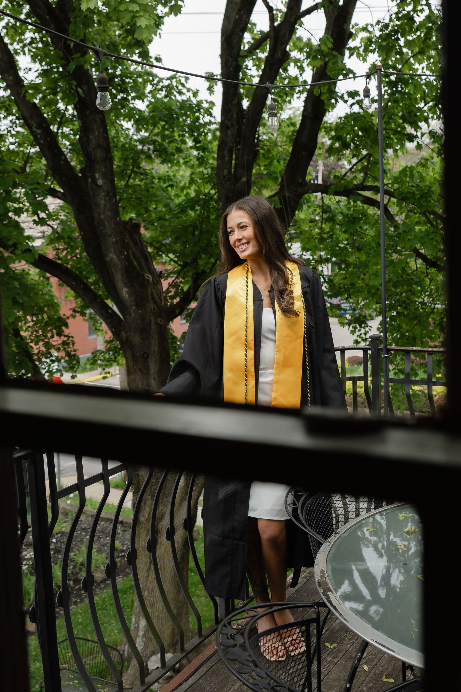 Smiling young woman in black graduation gown and gold stole standing on an outdoor balcony surrounded by green trees.