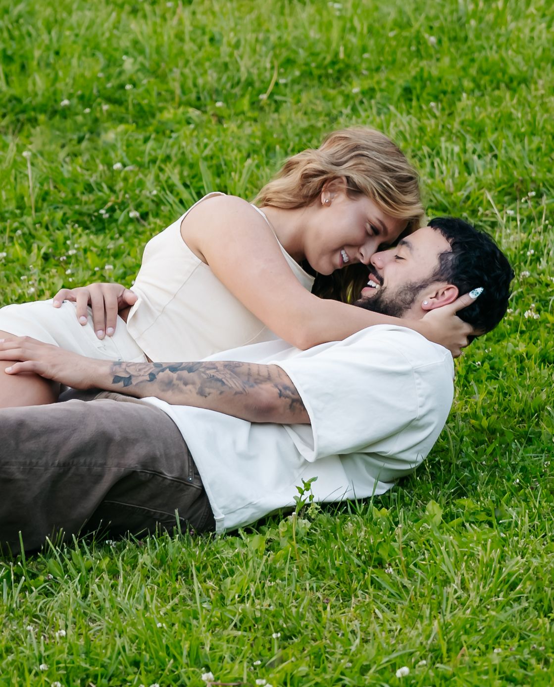 A couple laughs and cuddles together while lying on a lush green grass lawn on a sunny day.