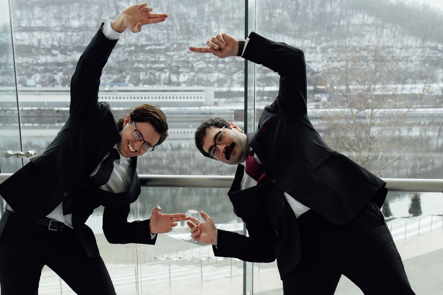 Two businessmen in suits striking fun dance poses near large windows with snowy landscape outside.