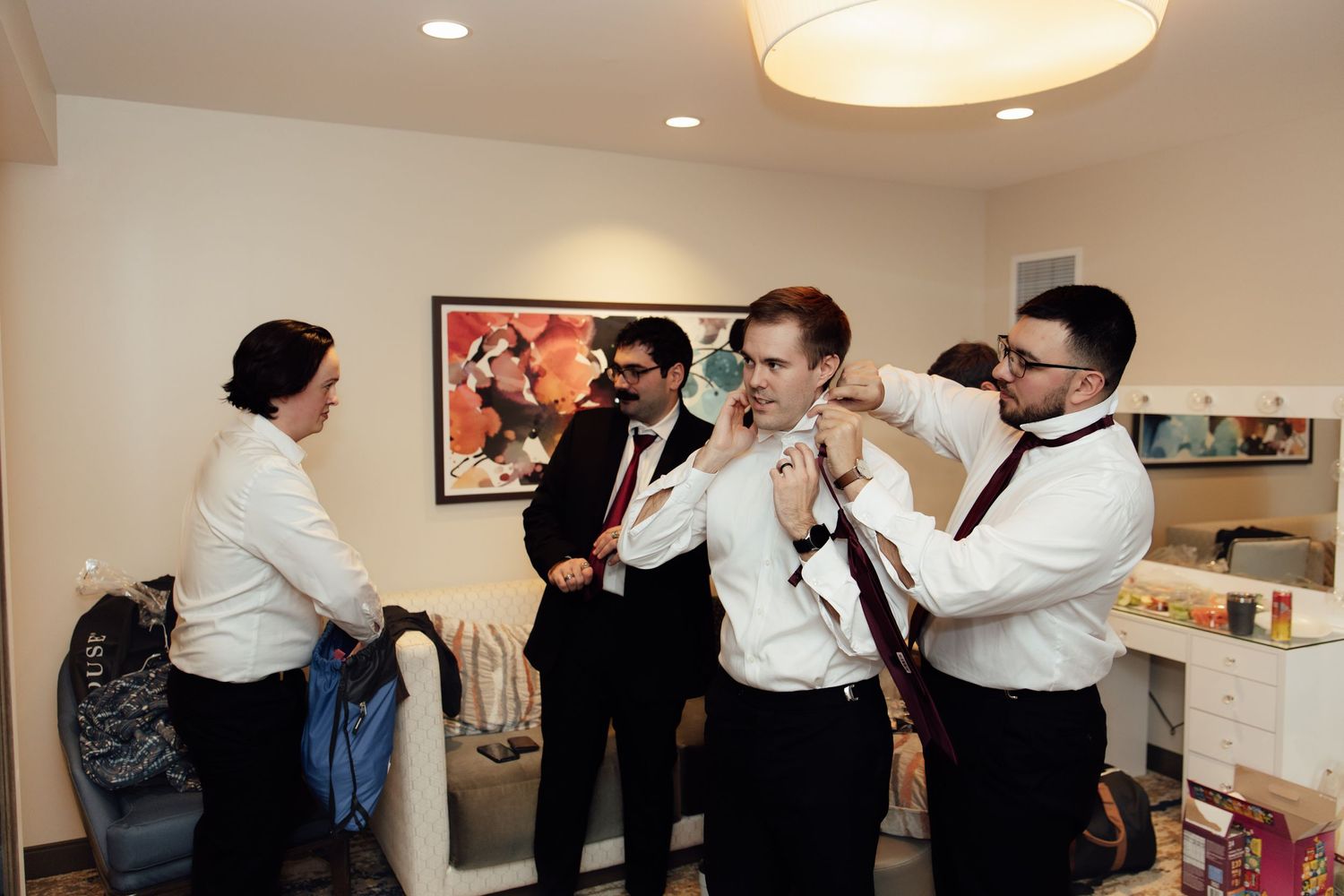Groomsmen helping groom adjust bow tie and shirt before wedding ceremony in elegant hotel room.