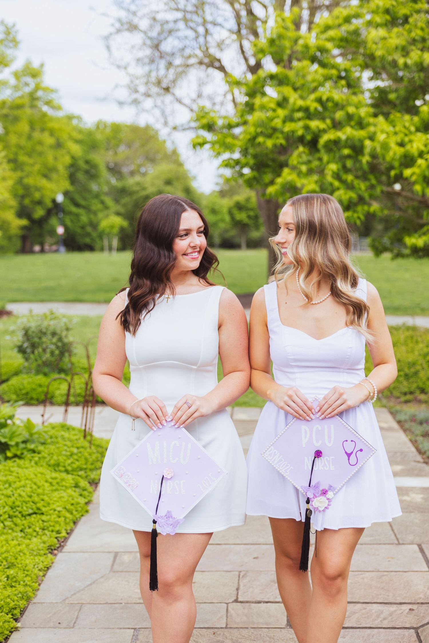 Two friends in white dresses stand together in a garden holding decorated graduation caps and smiling.