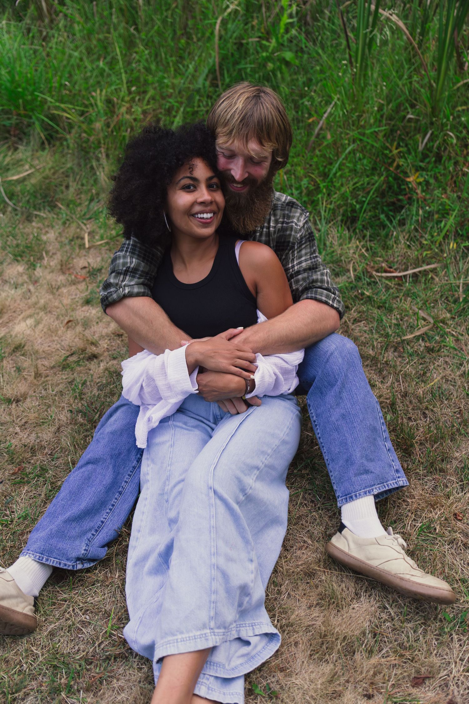 A happy interracial couple sitting together on grass, embracing and smiling outdoors in casual denim outfits.