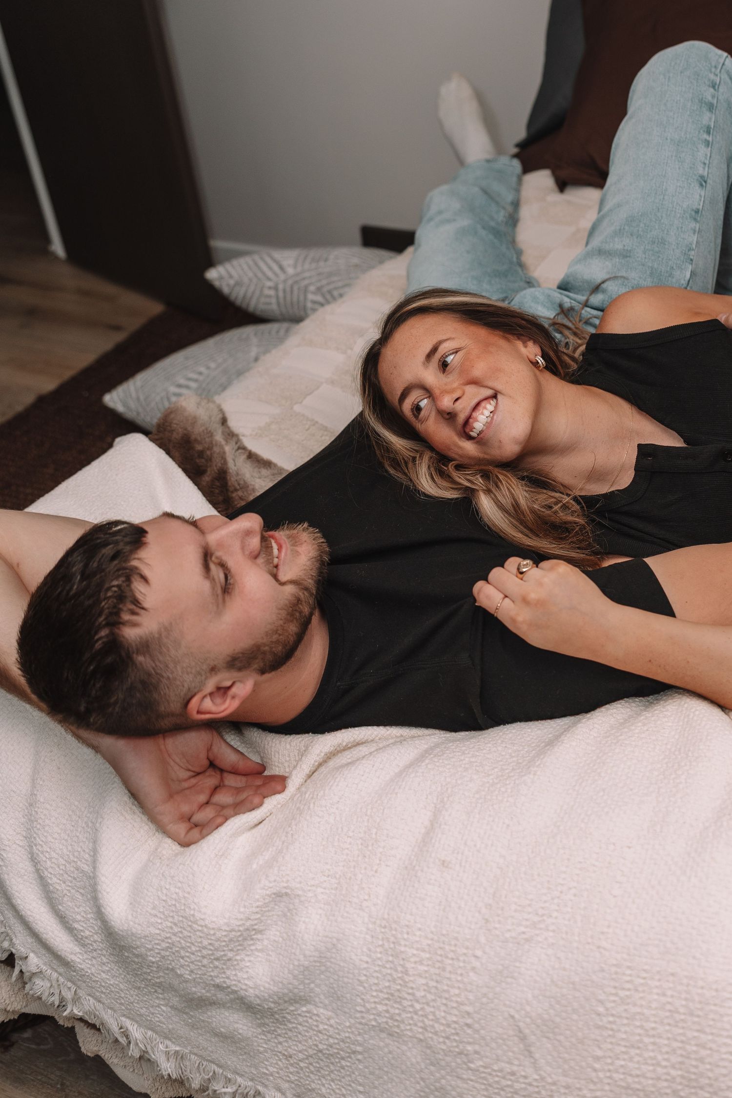 Couple in matching black shirts laughing and relaxing together on a bed with white blanket and pillows.