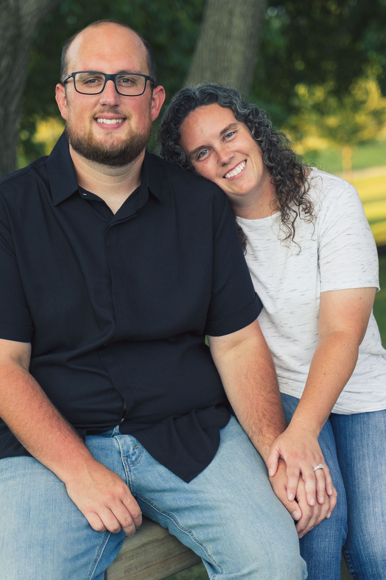 A smiling couple sitting together outdoors in a park, man wearing glasses and black shirt, woman in gray top.