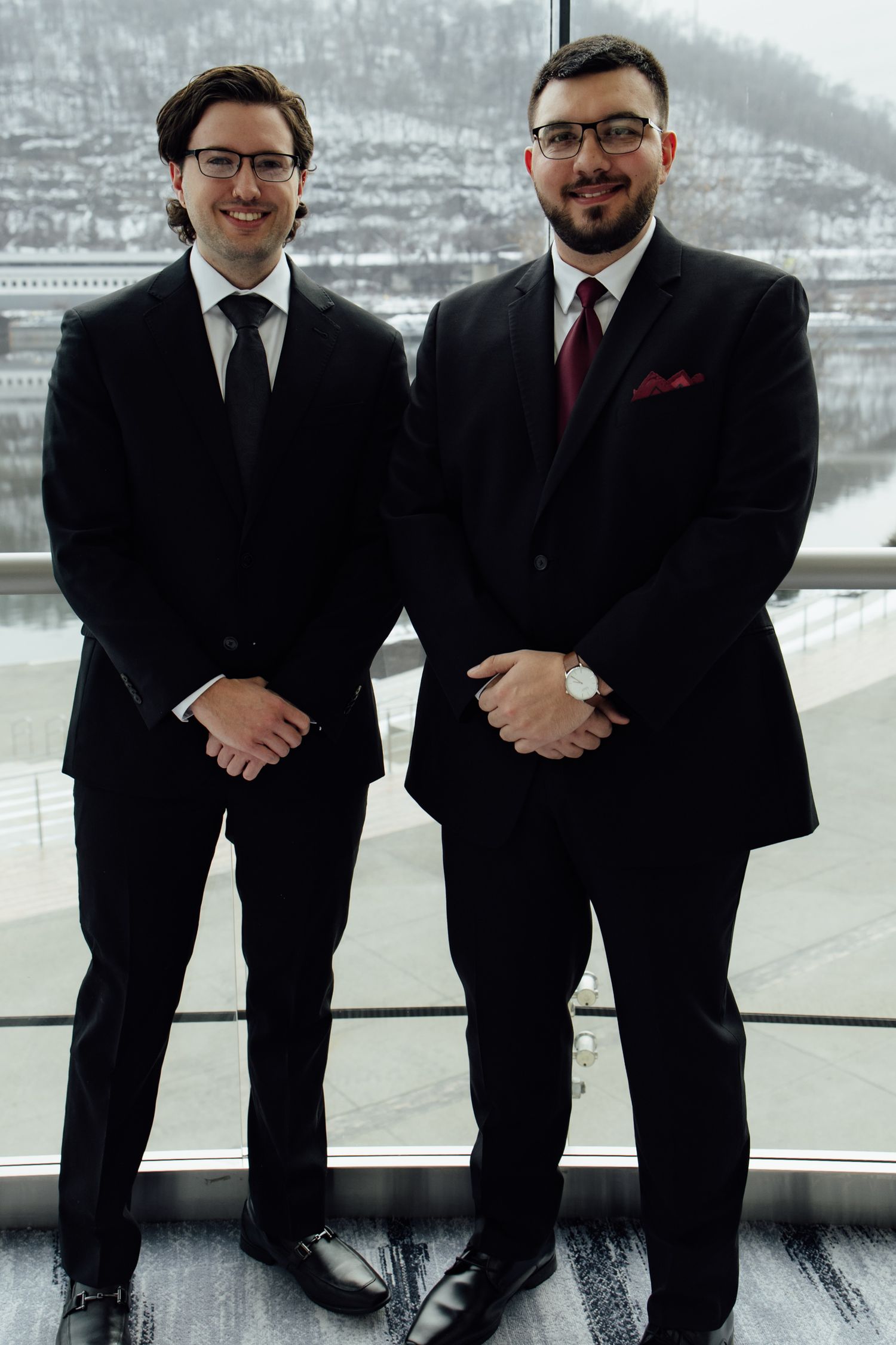 Two men in black suits and glasses smiling indoors with a snowy cityscape visible through large windows.