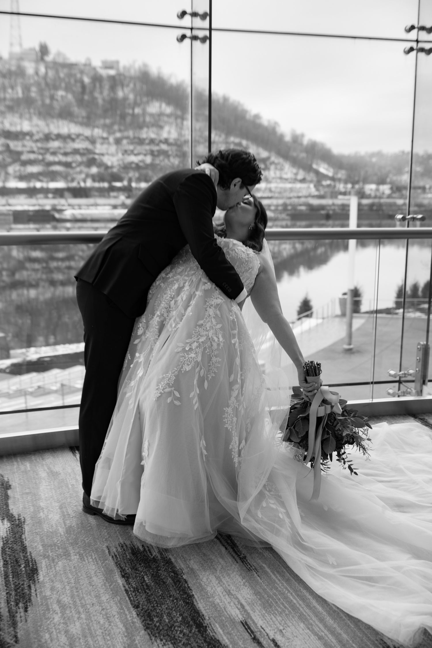 Black and white photo of a bride and groom sharing a romantic kiss near large windows overlooking a snowy river.