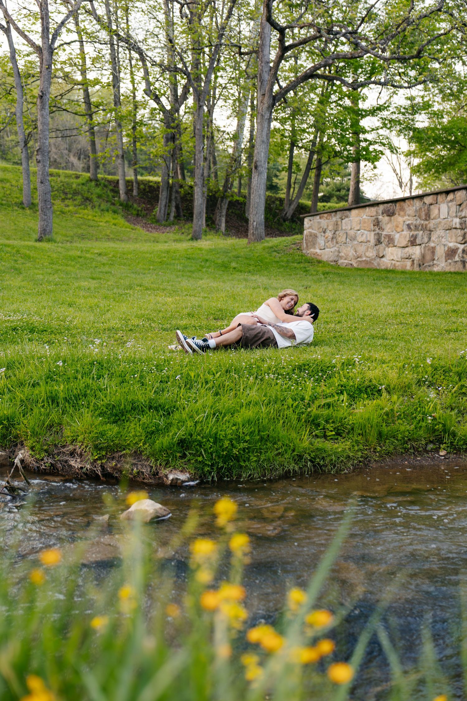 Couple relaxing together on green grass beside a peaceful stream in a lush park with trees and stone wall.