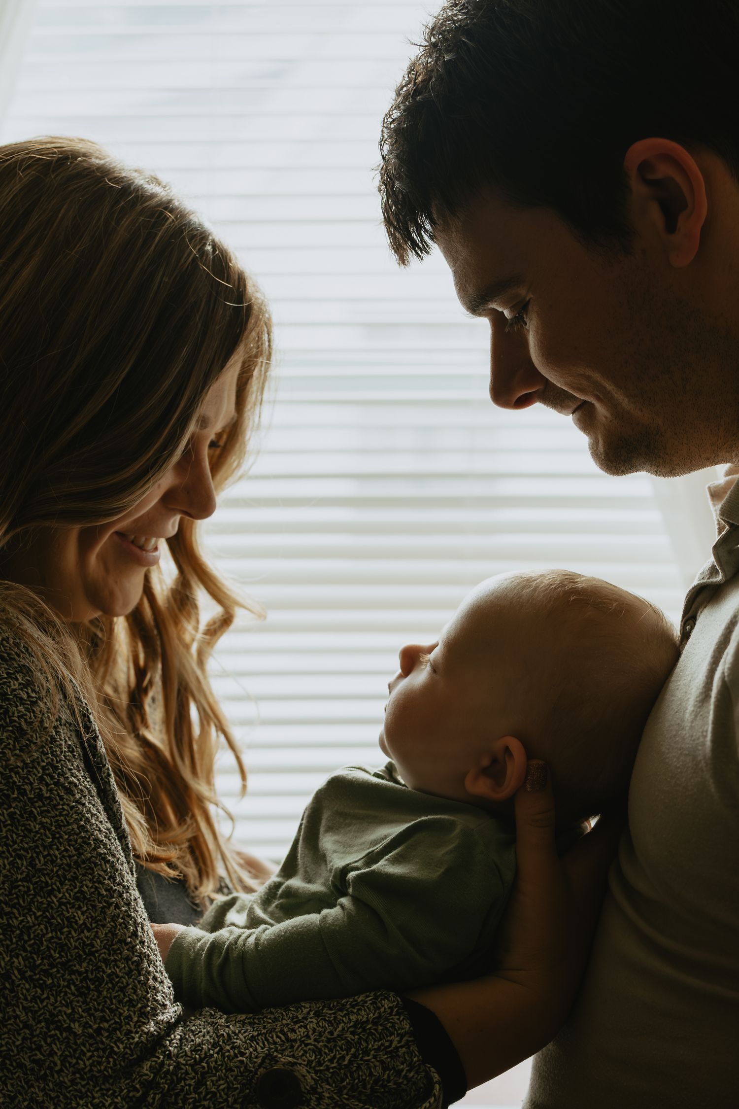 A young couple smiling lovingly at their newborn baby held between them near a window with blinds.