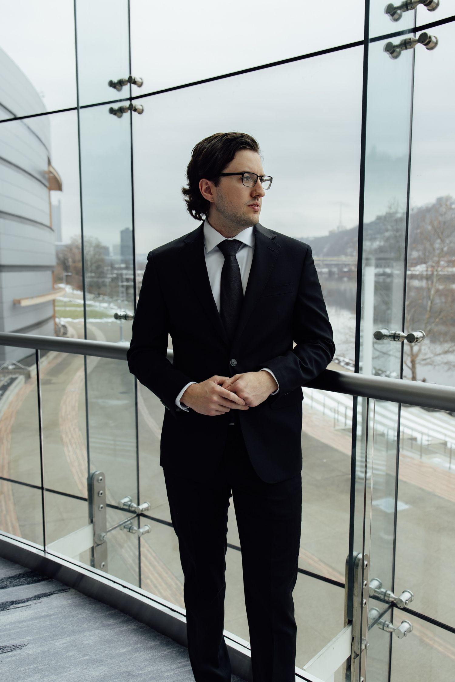 A professional businessman in a black suit and tie stands on a glass-railed balcony, gazing into the distance.