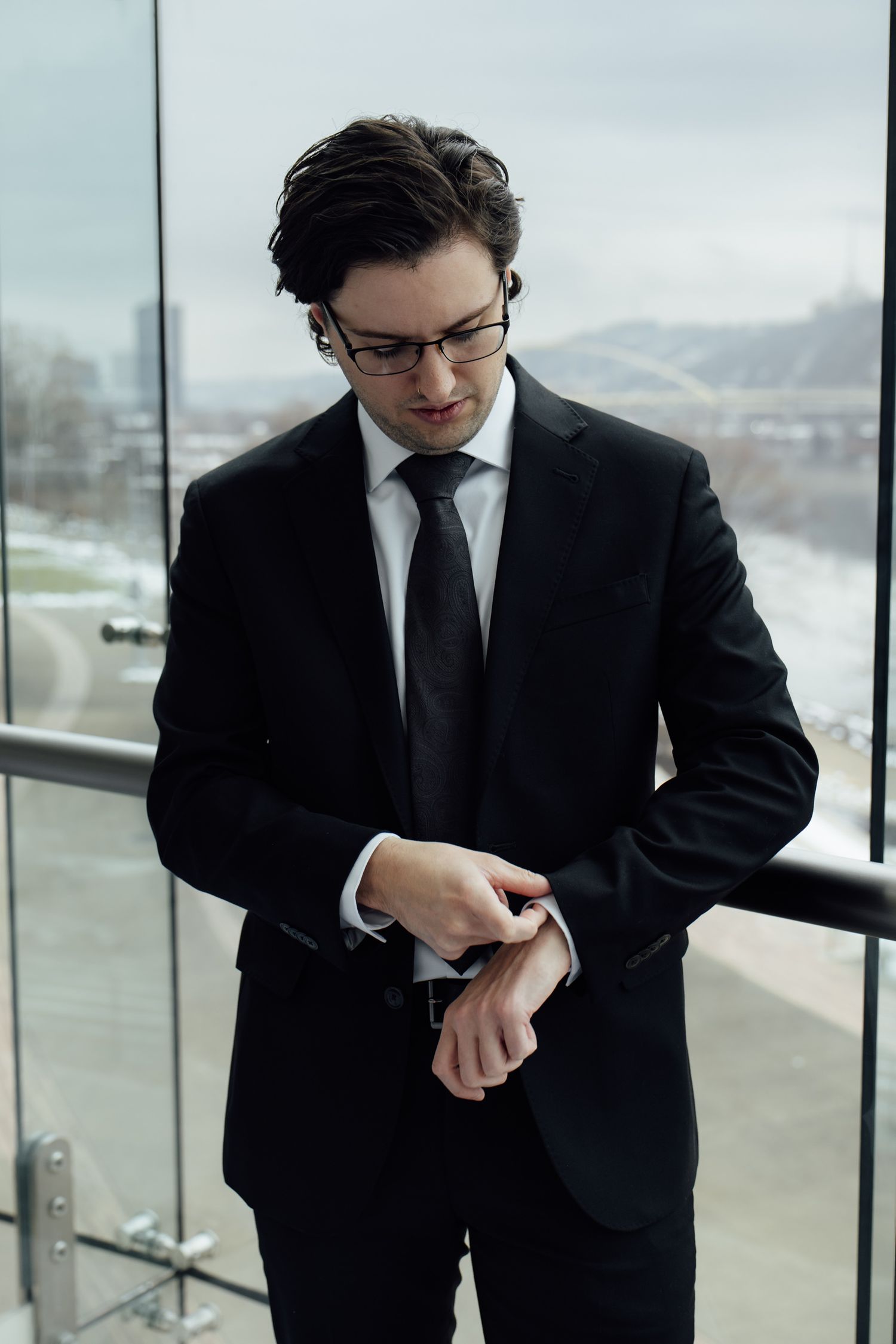 A businessman in a black suit and glasses checks his wristwatch while standing near a glass window.