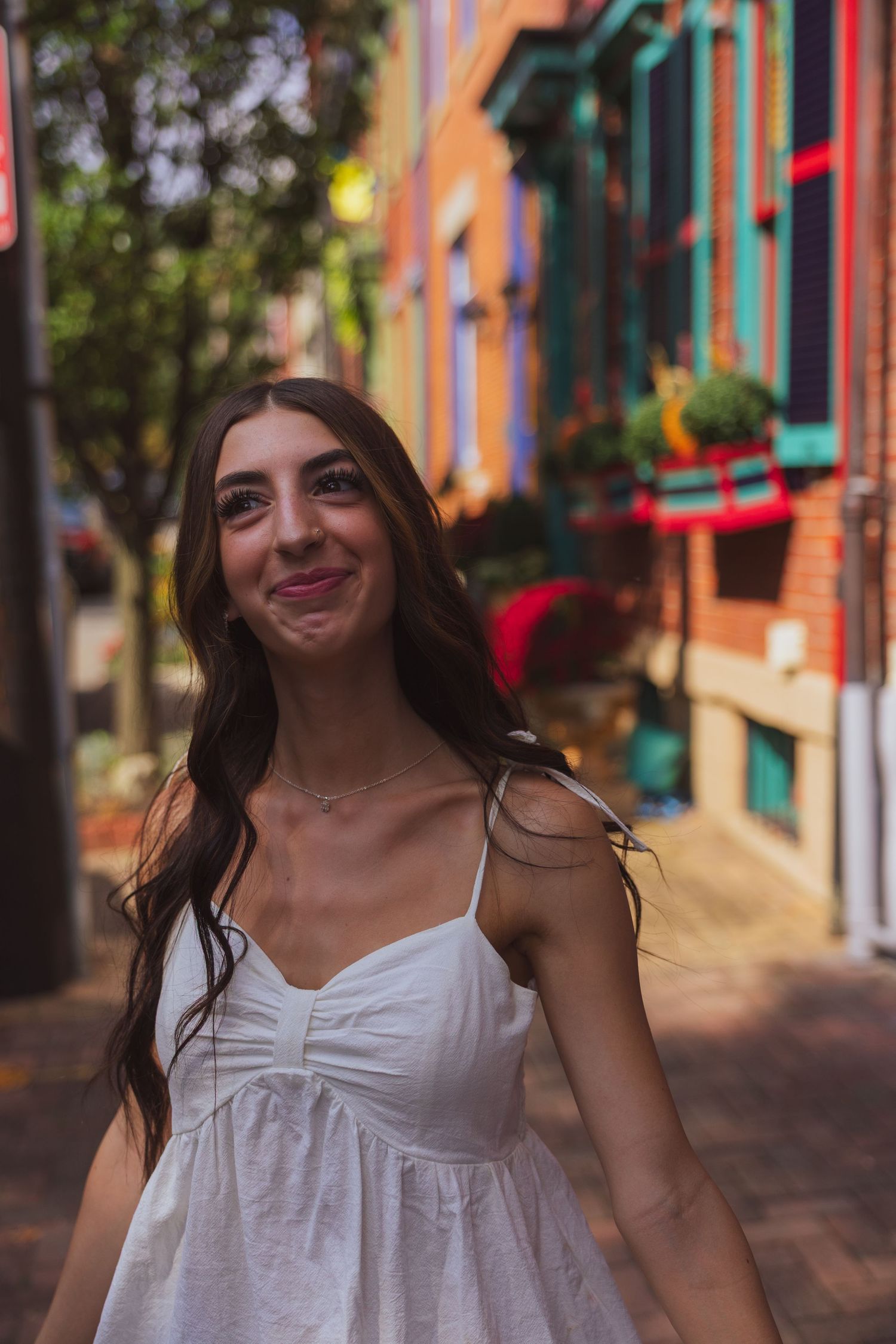 Young woman in white sundress smiling on colorful urban street with vibrant building facades in background.