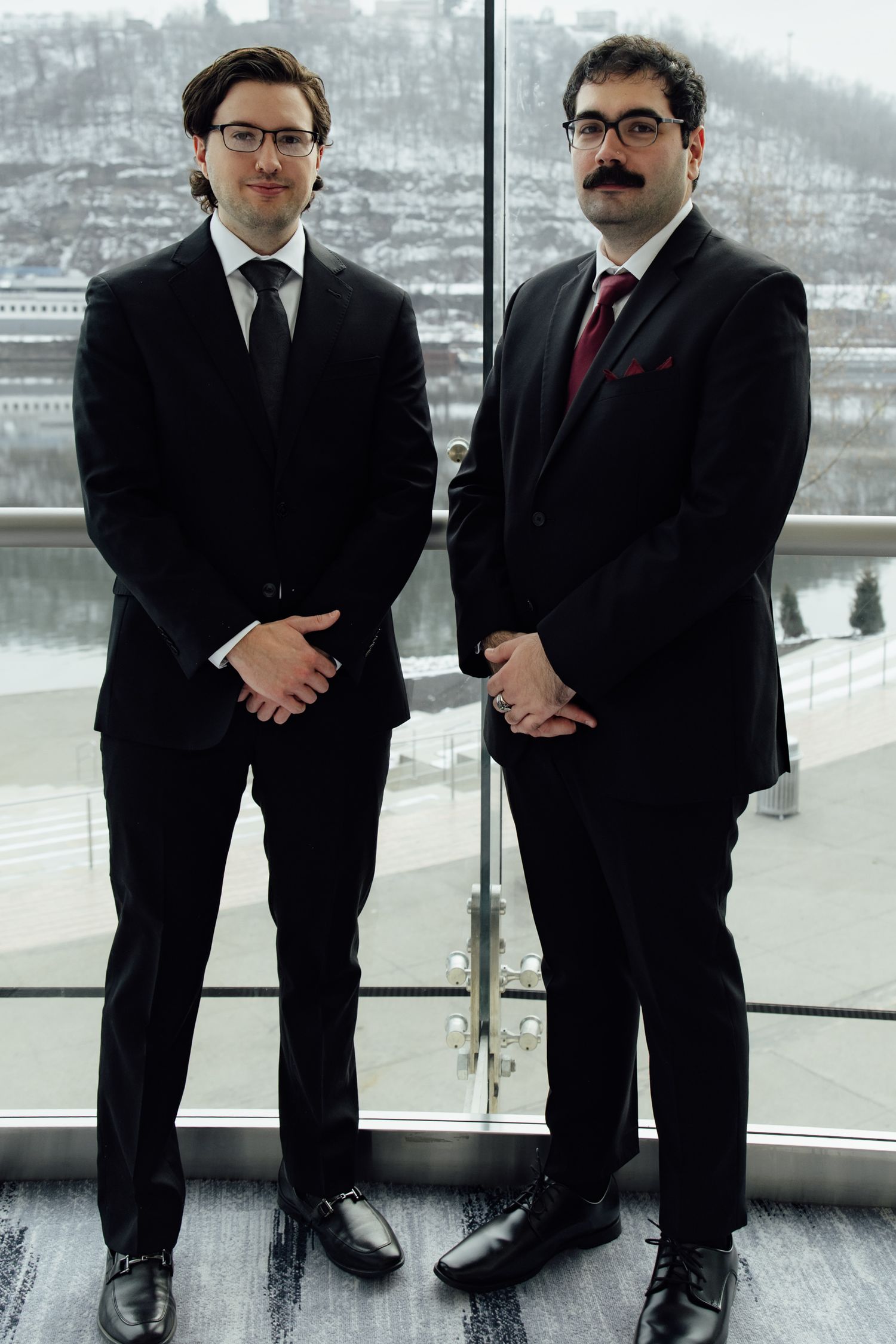 Two professional men in black suits standing indoors near glass windows with a snowy cityscape backdrop.