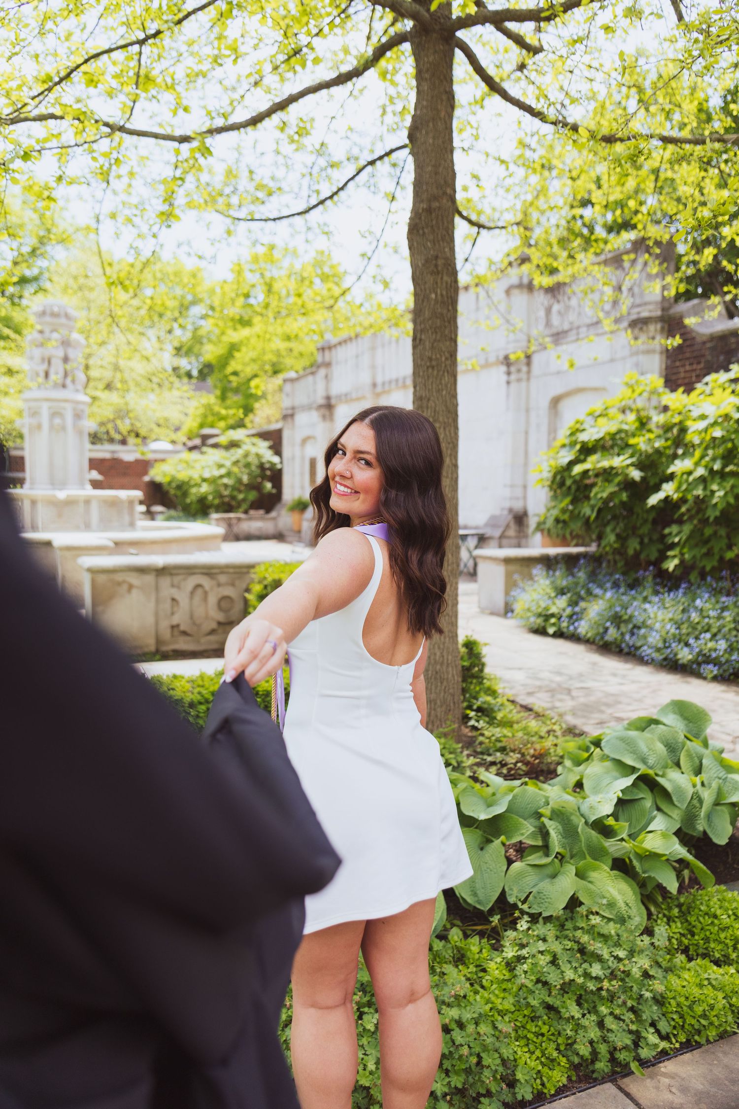 A person in a white summer dress poses outdoors near stone architecture and lush greenery on a bright sunny day.