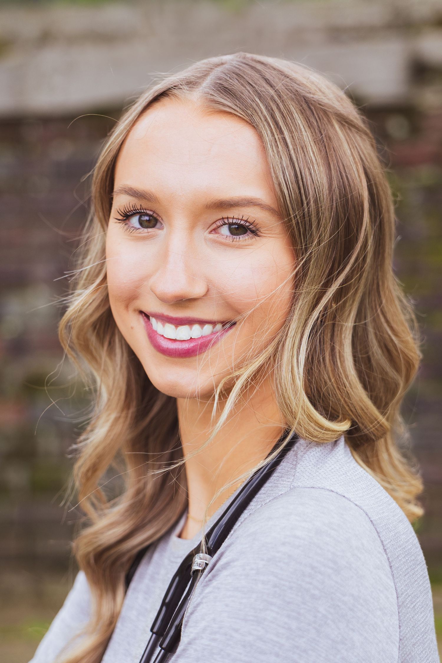 Portrait of person with wavy blonde hair wearing a gray top and warm smile against a stone wall backdrop.