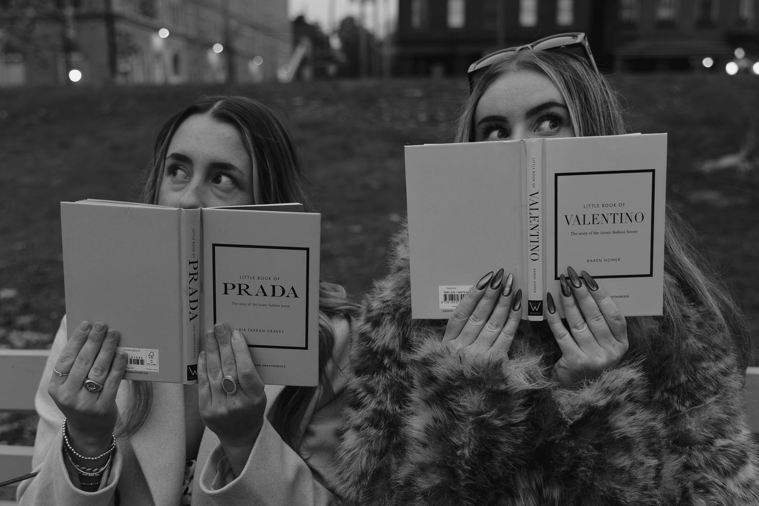 Two women holding Prada and Valentino fashion books covering their faces in a black and white photo.