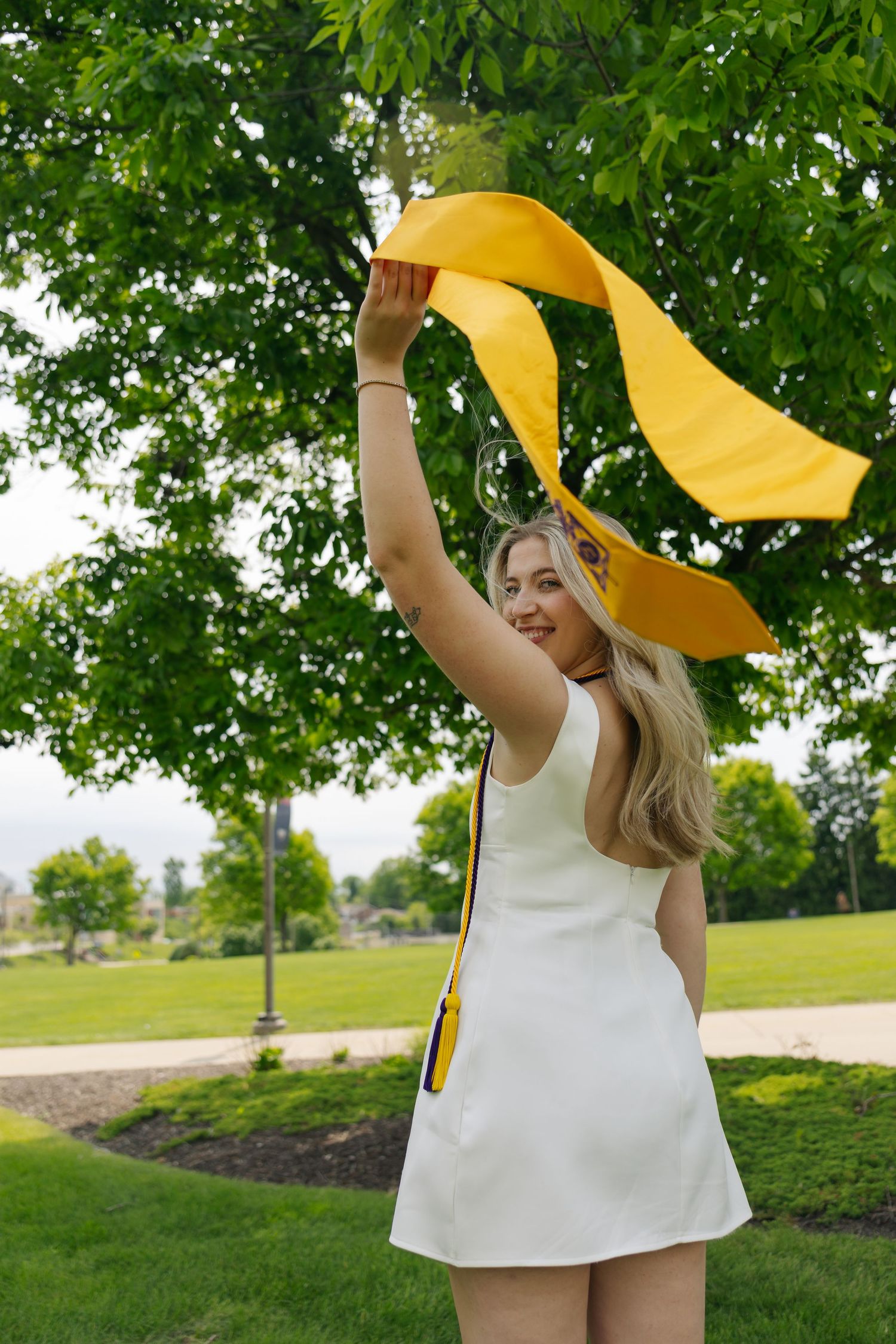 Young woman in white dress waving yellow fabric in a lush green park on a bright summer day.