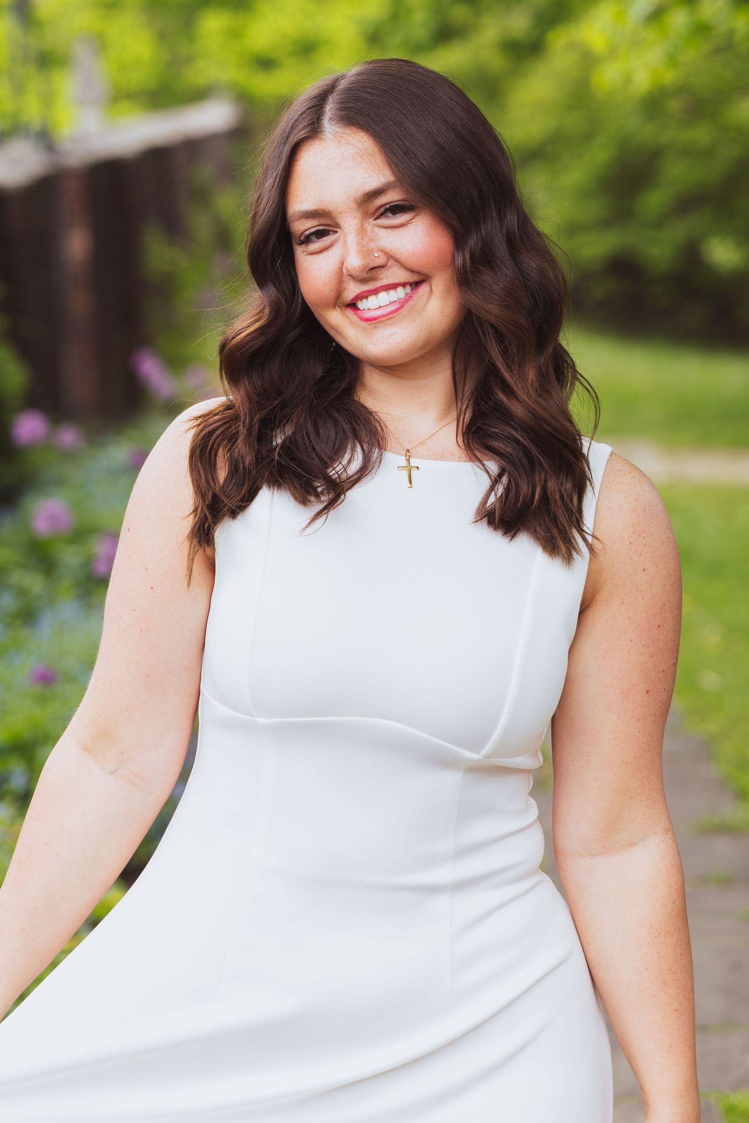 Woman in white sleeveless dress smiling while standing in a garden with blooming flowers in background.