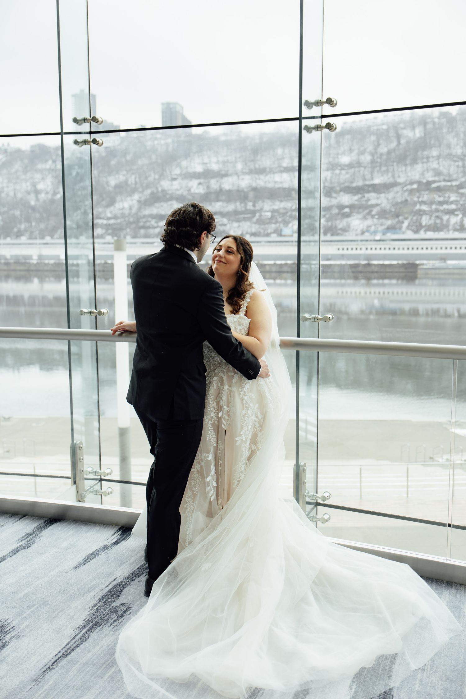 Bride and groom share a romantic moment by floor-to-ceiling windows with a snowy river view on their wedding day.