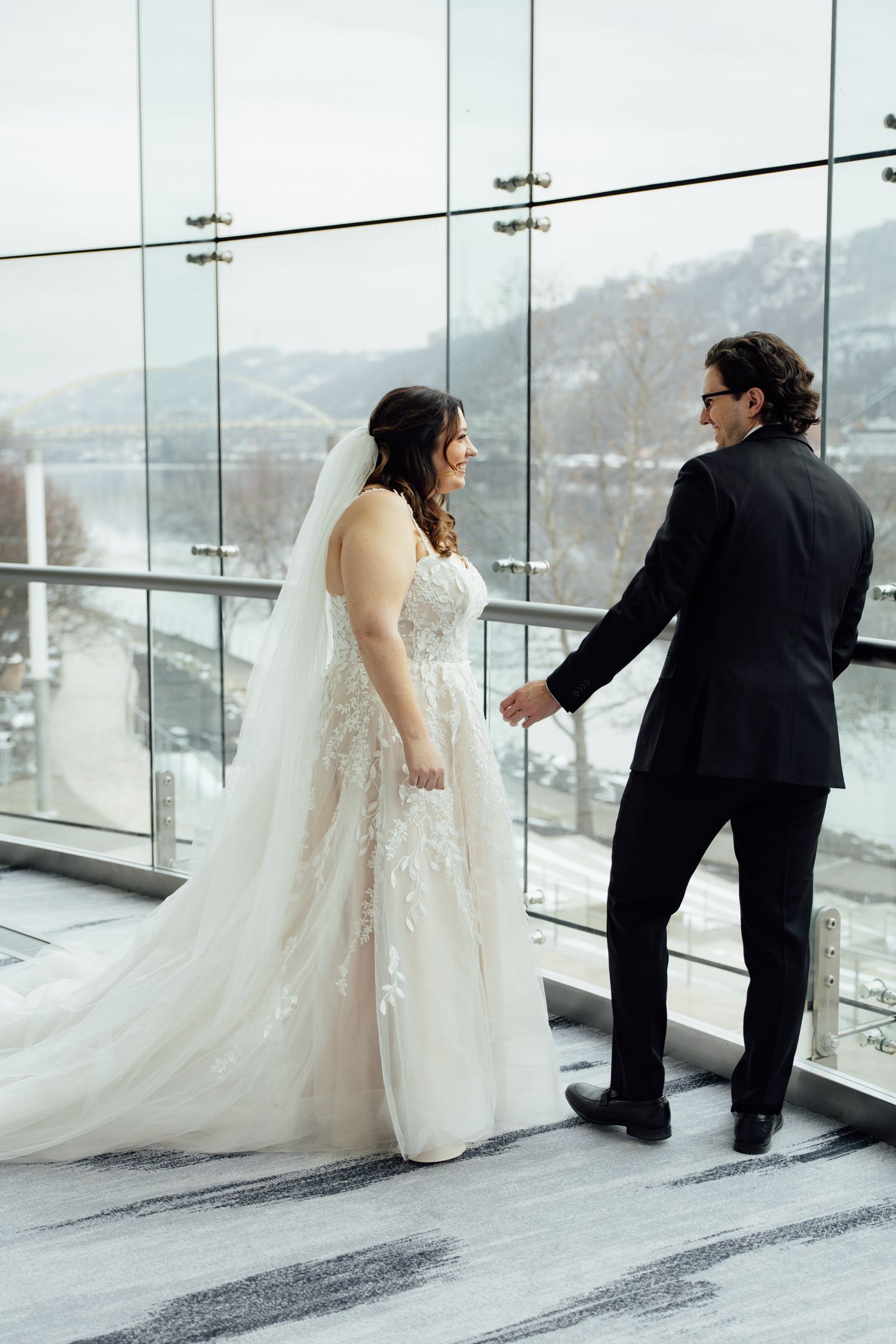Bride in lace gown and veil holds grooms hand near glass windows with snowy river view during winter wedding.
