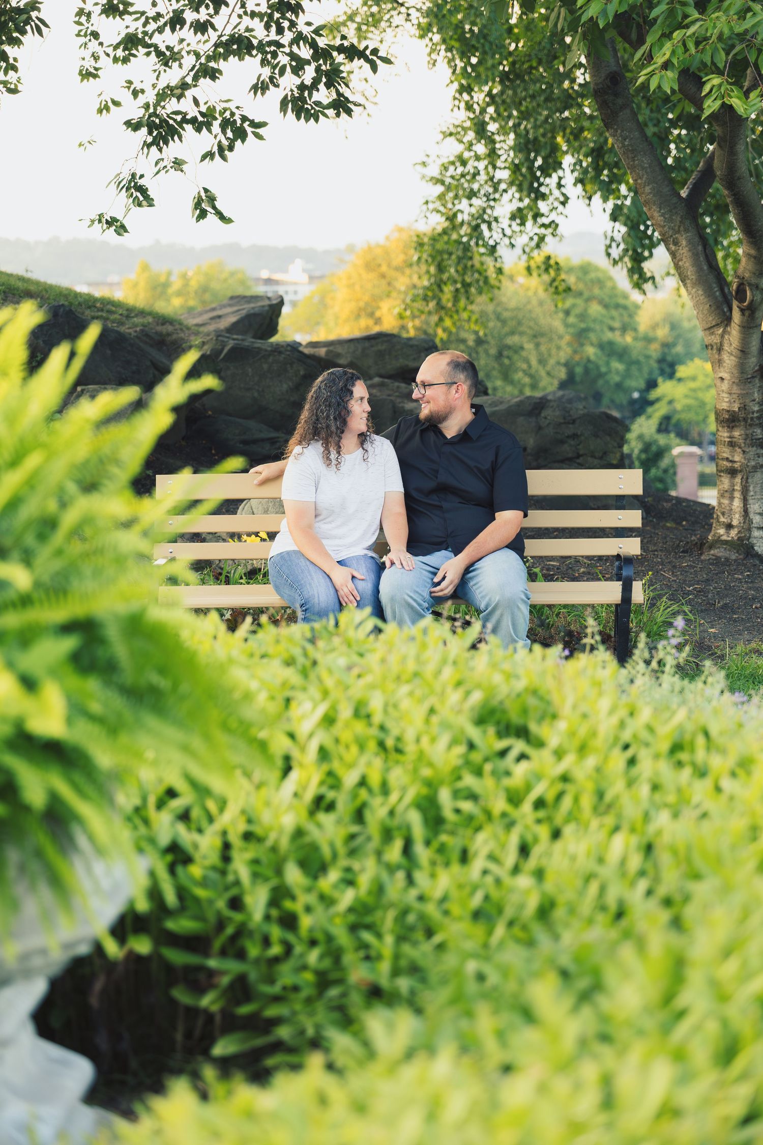 A happy couple sitting close together on a wooden bench surrounded by manicured hedges and trees.