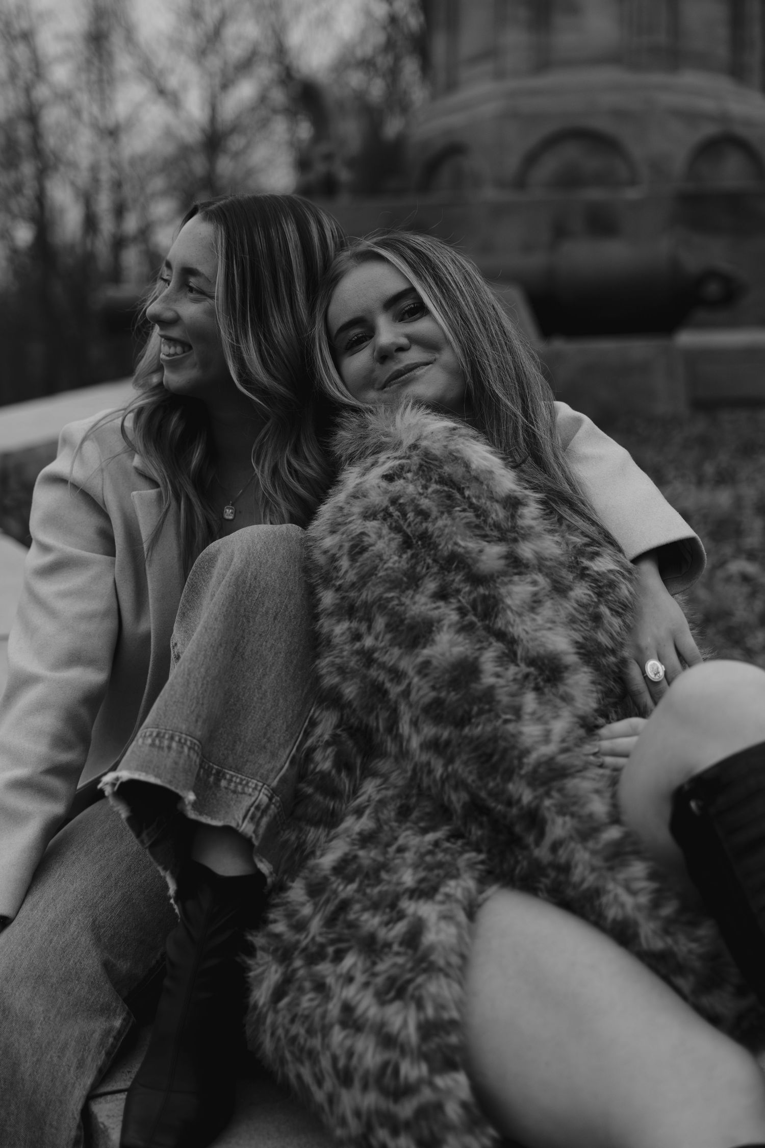 Two young women sitting outdoors, one leaning on the other, smiling, in a black and white photo.