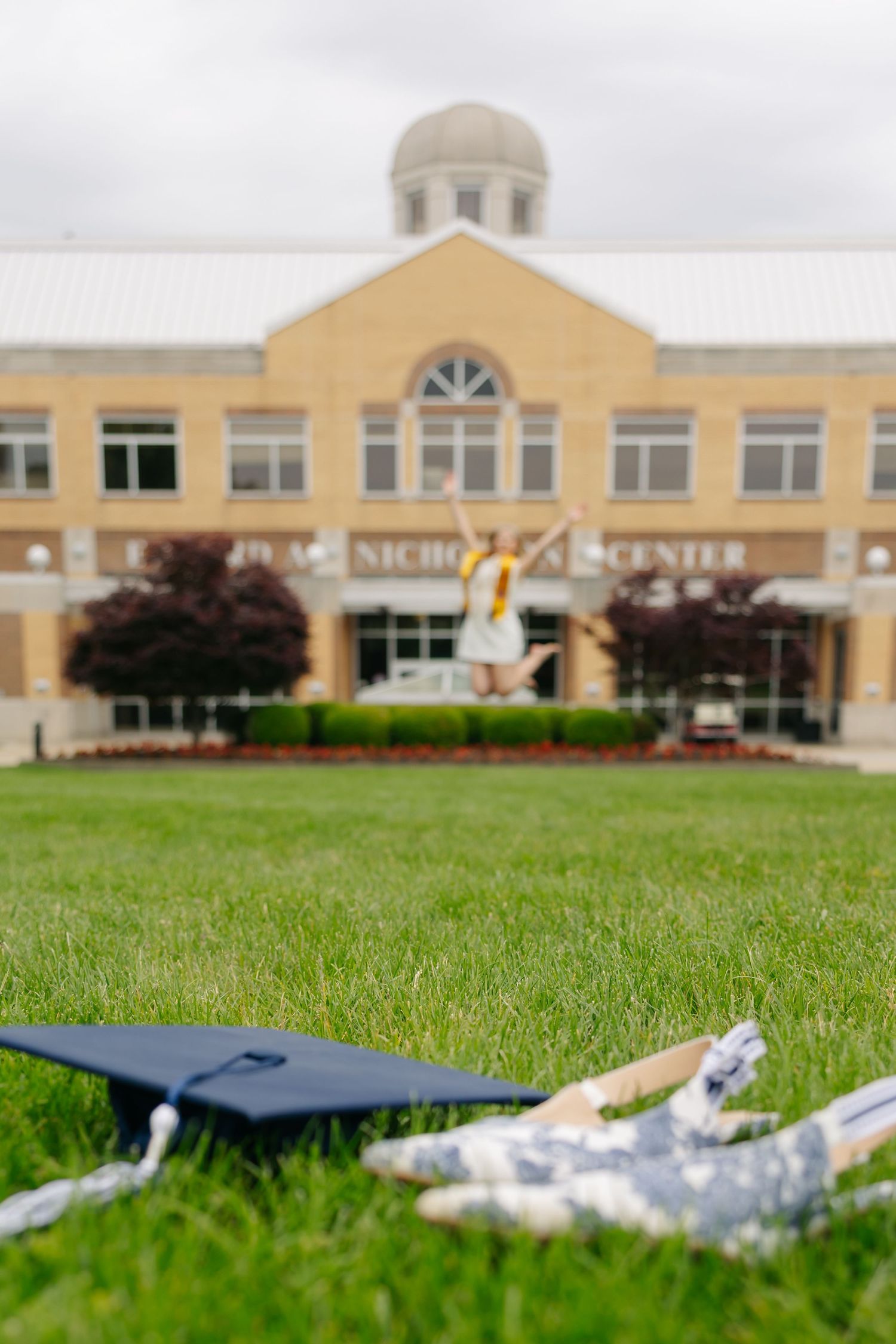 Graduation cap and diploma on green grass with a student jumping joyfully in front of a university building.