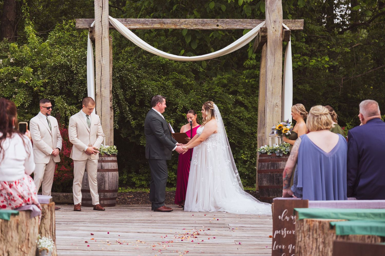 Couple exchanges vows at rustic outdoor wedding ceremony with wooden arch and barrel decorations.