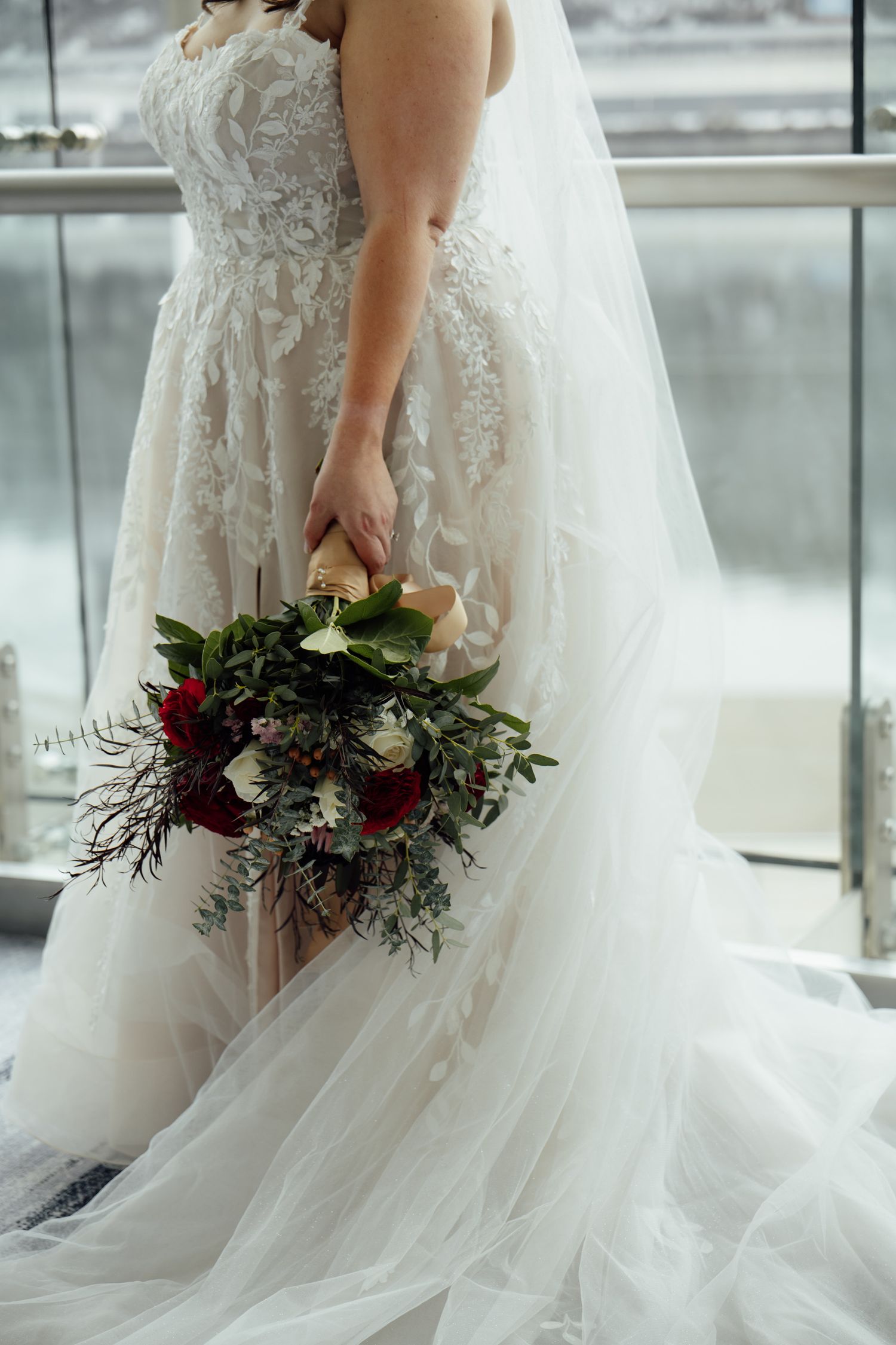 Bride in lace wedding gown holding cascading bouquet of red roses and greenery near window.