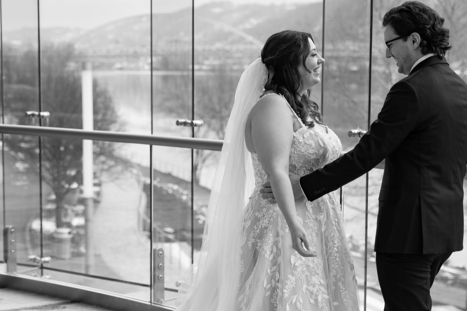 Black and white photo of a bride and groom sharing a first look moment by large windows with a scenic view.