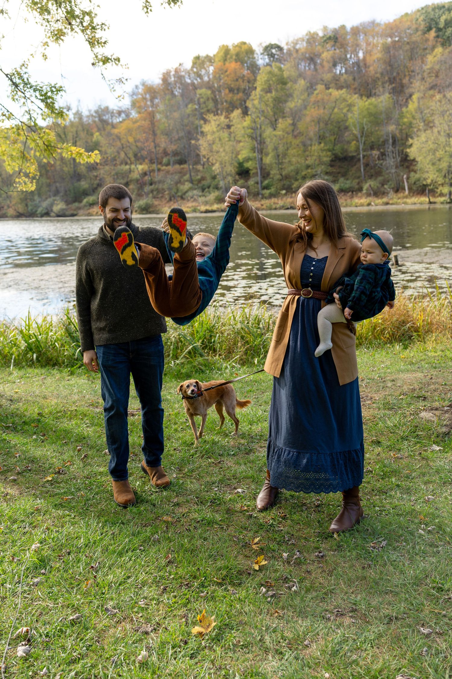 Family with two babies and a dog enjoying an autumn day by a scenic lake surrounded by colorful fall foliage.
