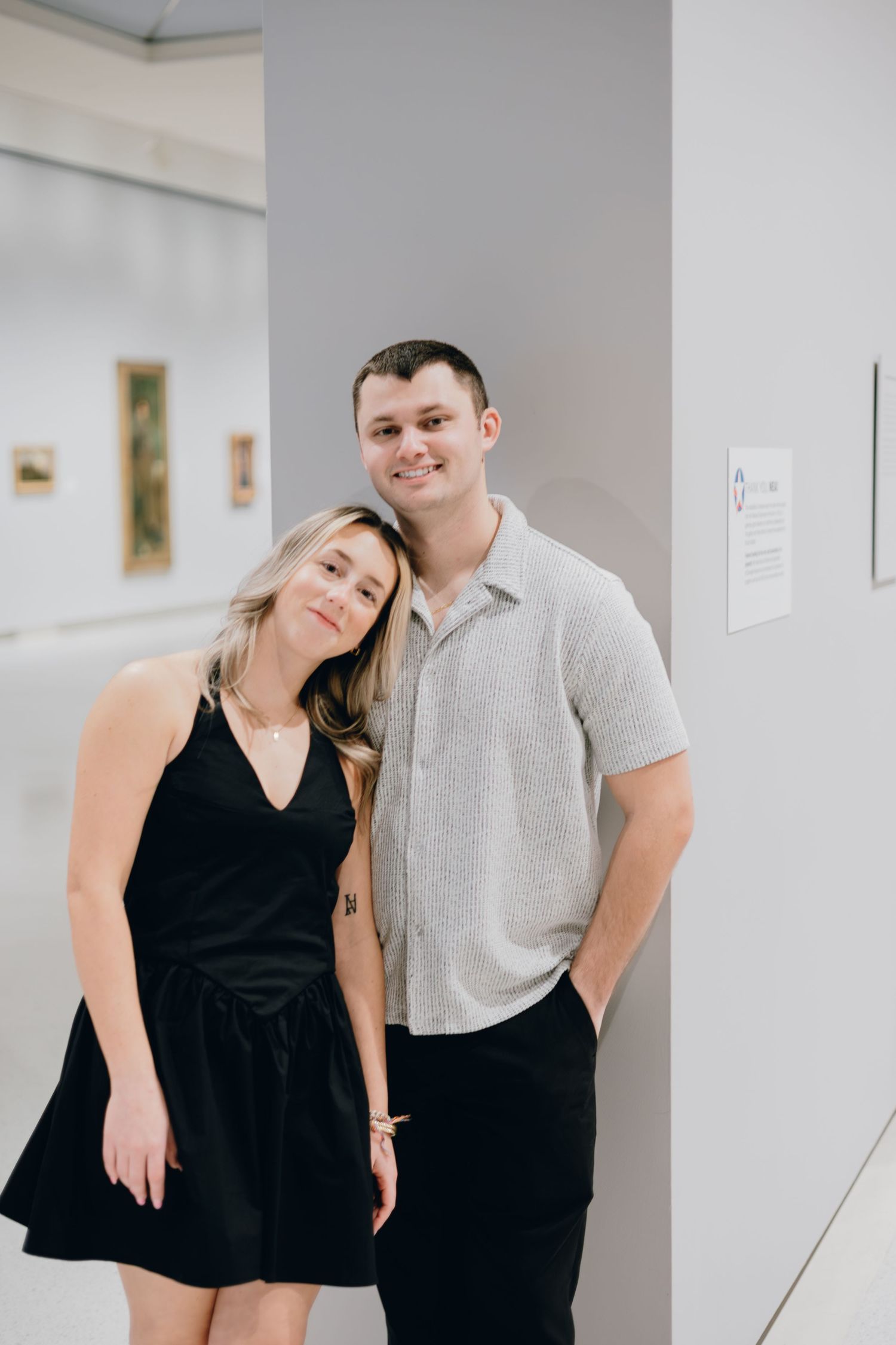 Casual portrait of couple leaning against gallery wall in coordinated outfits.
