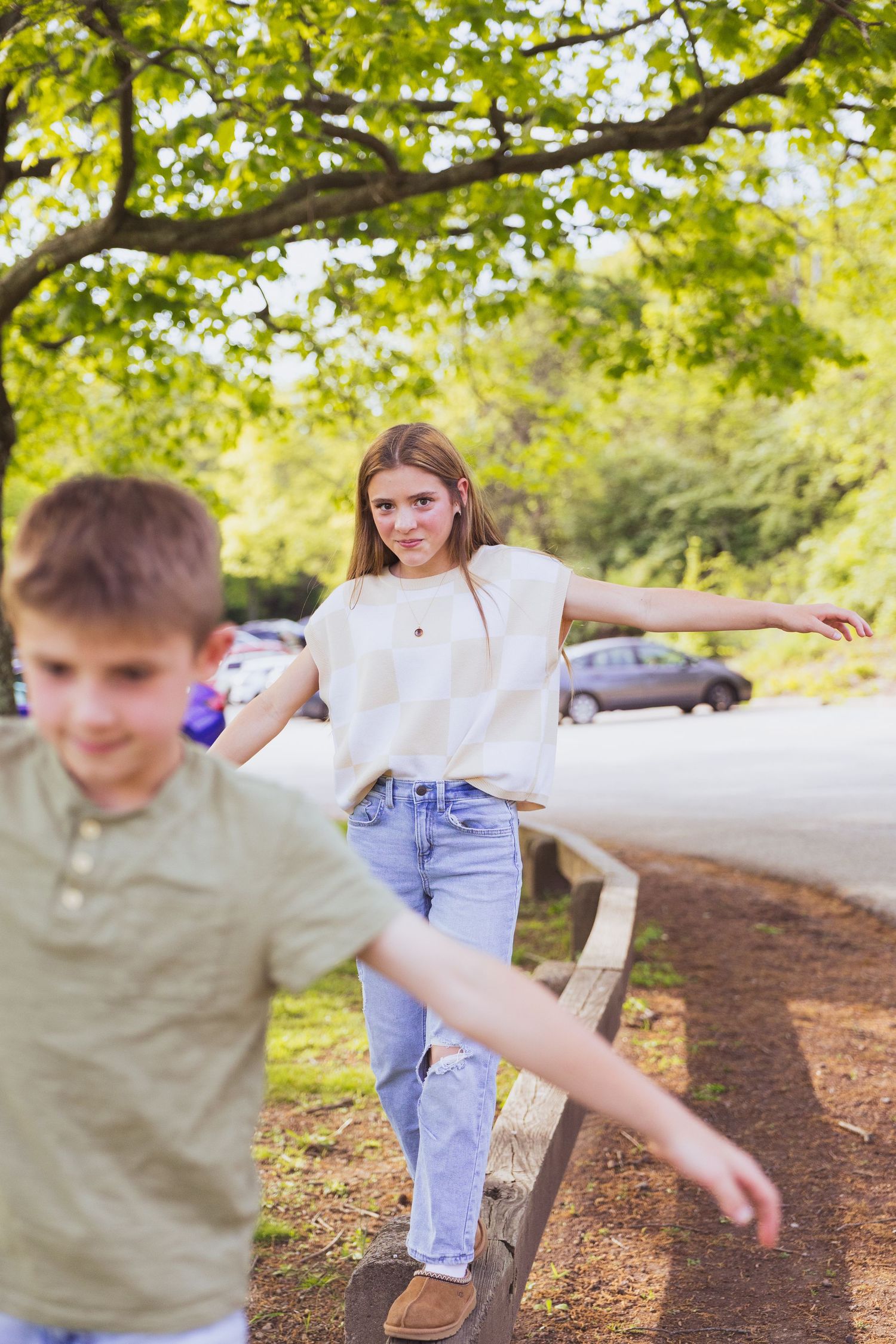 Children playing and balancing on logs along a wooded path on a sunny day, surrounded by green trees.