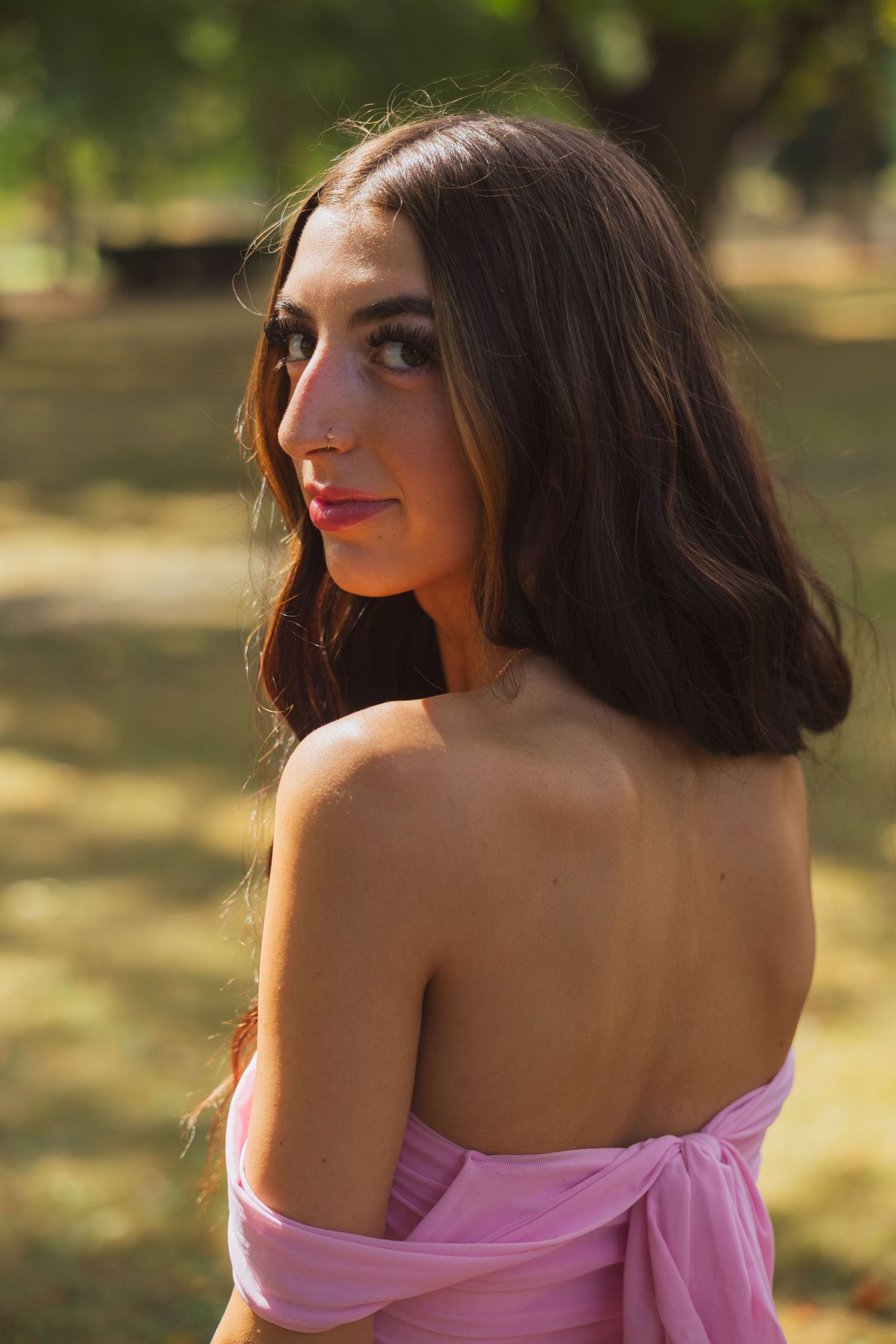 Young woman in a pink off-shoulder dress poses outdoors in a sunlit park with green trees in the background.
