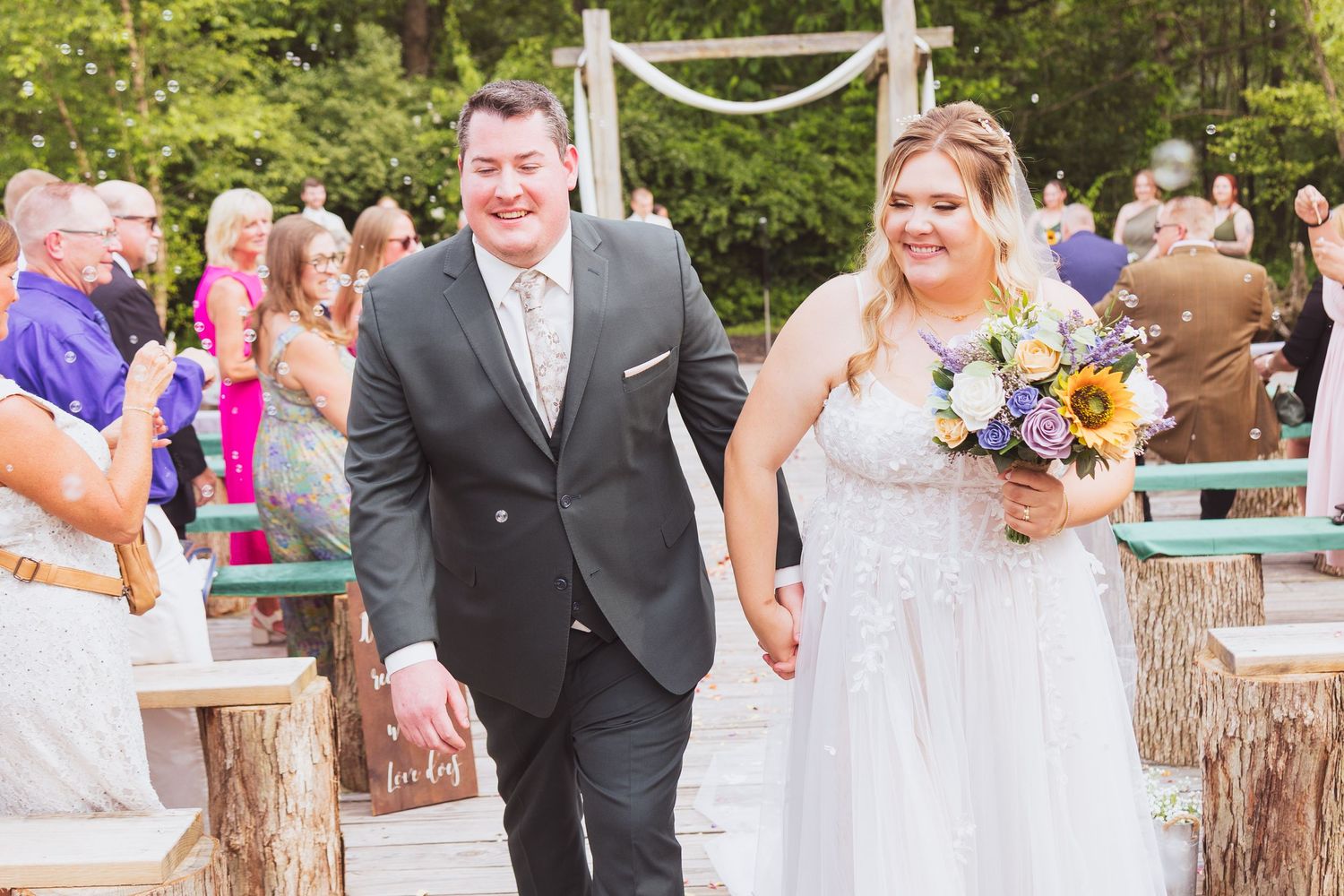 Wedding party walking down aisle with sunflowers at outdoor rustic ceremony venue.