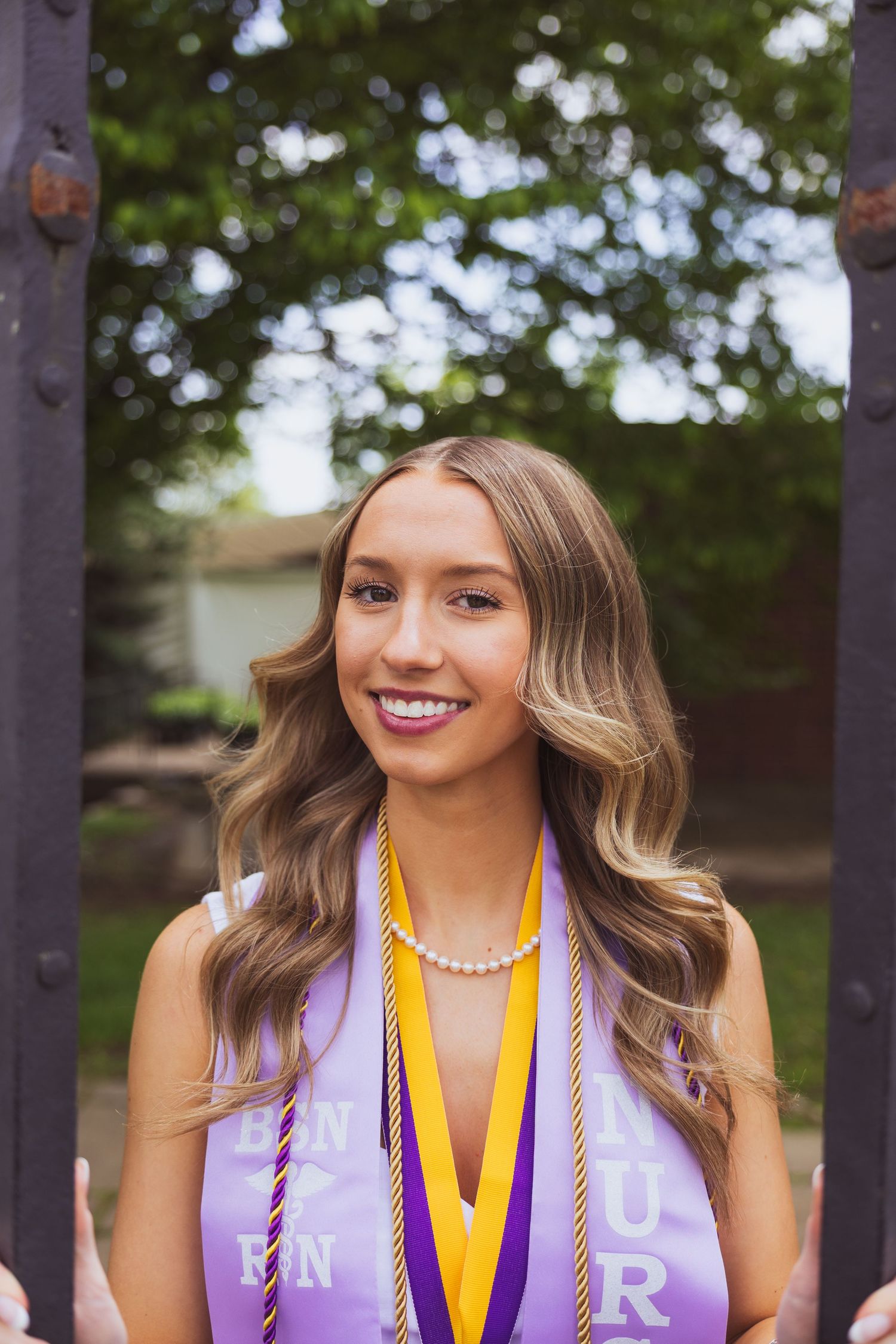 Graduate wearing purple and yellow honor cords with a white graduation stole and pearl necklace smiles outdoors.