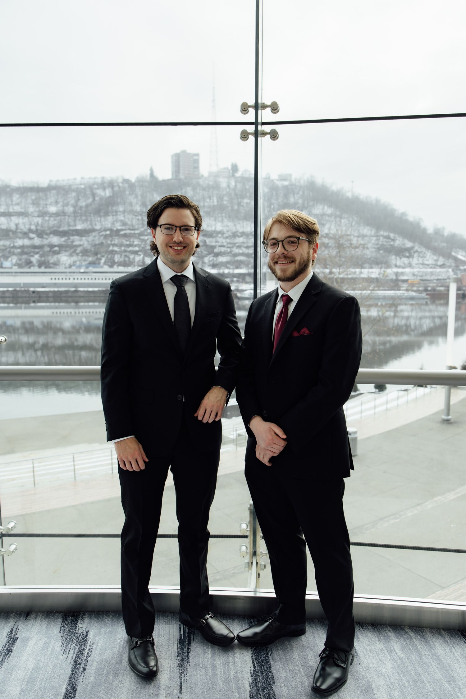 Two men in black suits and glasses smiling indoors with a snowy river valley visible through large windows.
