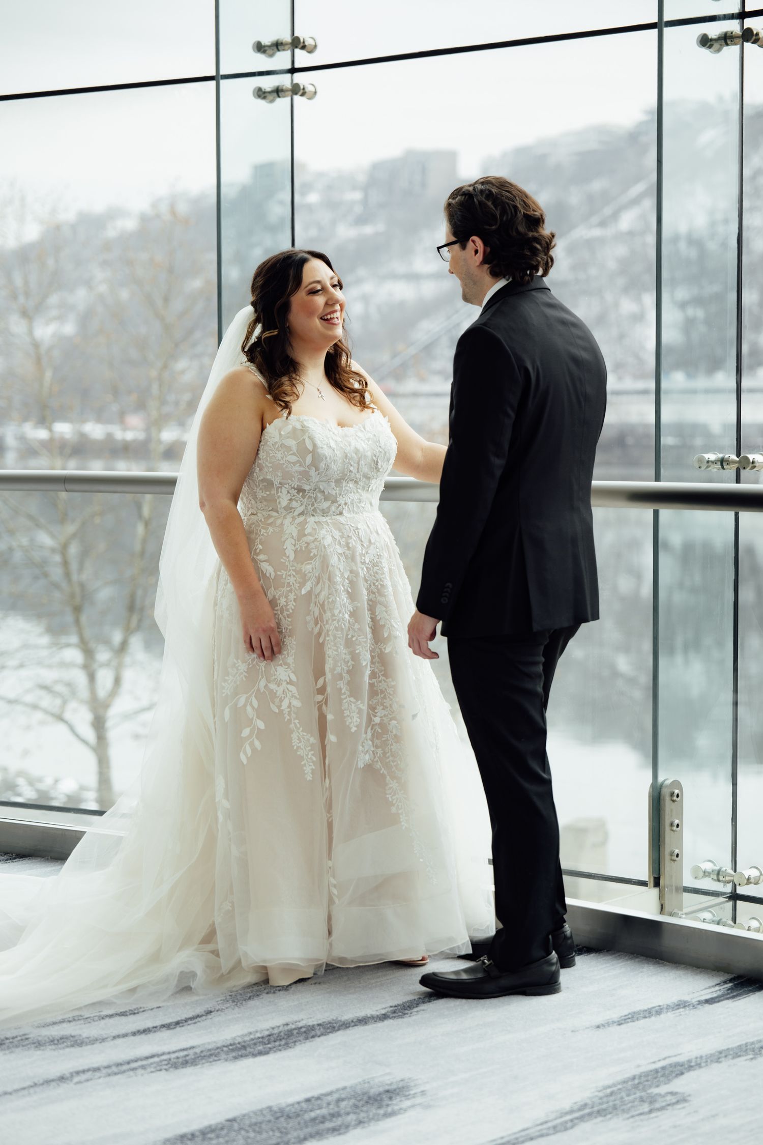 Smiling bride in floral lace gown faces groom in black suit by snowy river view window.