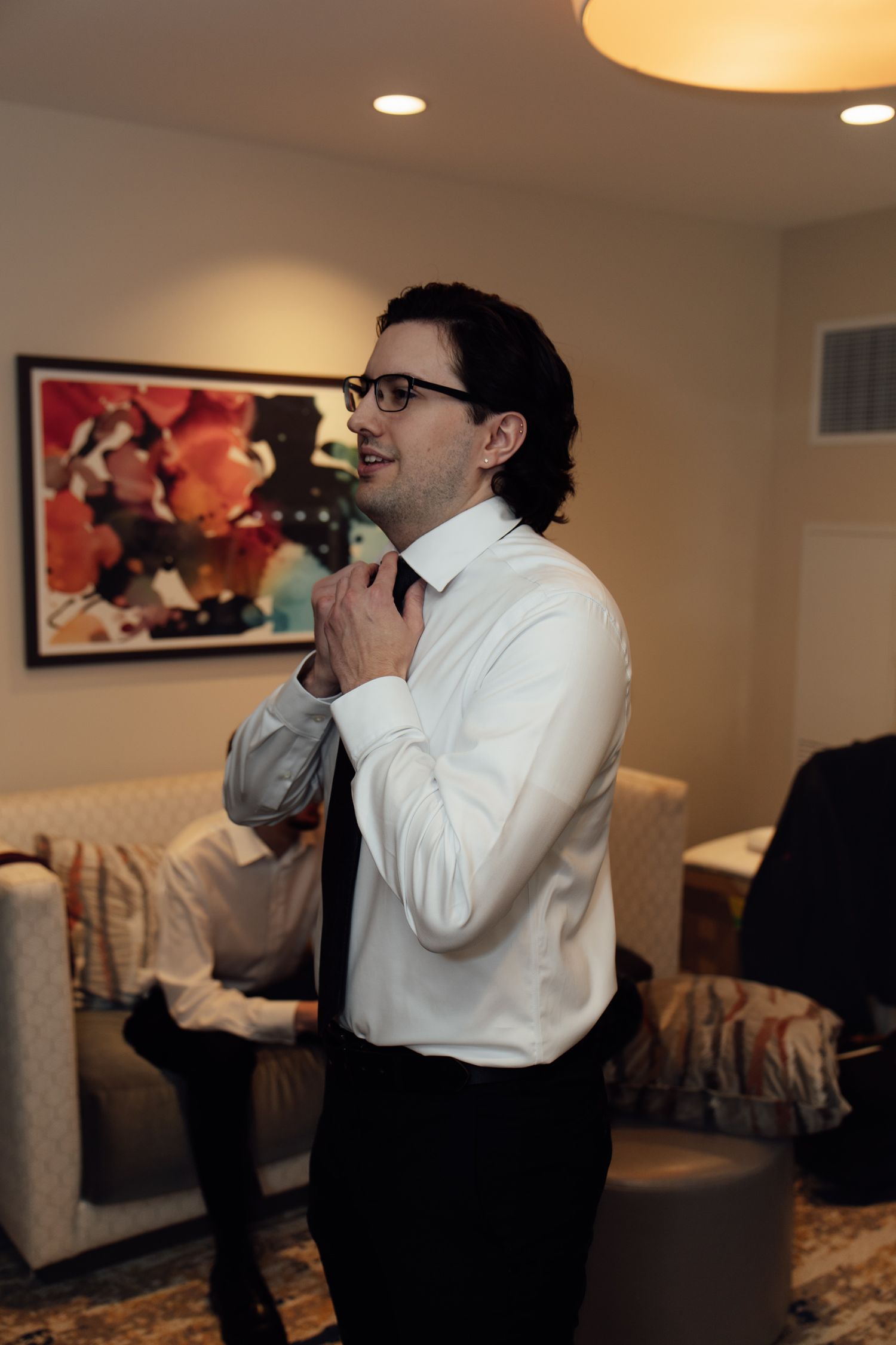 Groom in white shirt tying black necktie while preparing for wedding ceremony in modern living room.