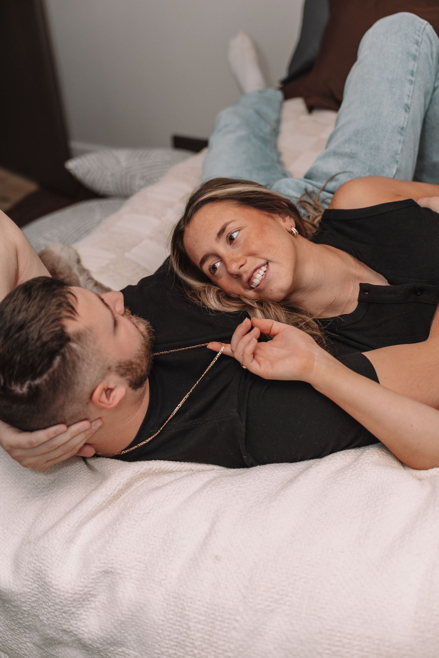 Couple lying on bed smiling at each other, woman in black top and jeans playfully touching man's necklace.