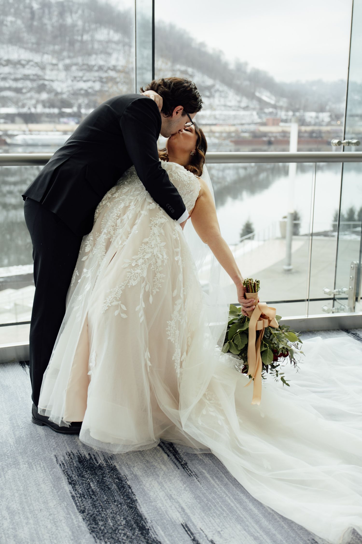 Couple sharing a tender kiss in a stunning lace wedding gown with bouquet, snowy river landscape visible through glass windows.