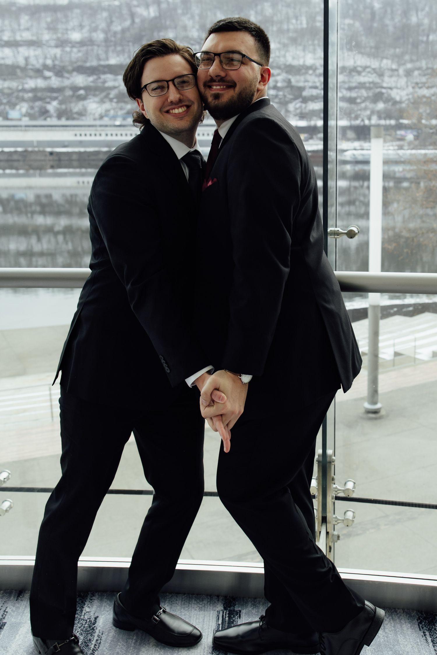 Two men in black suits holding hands and smiling on a glass balcony, posing for a formal couples portrait.